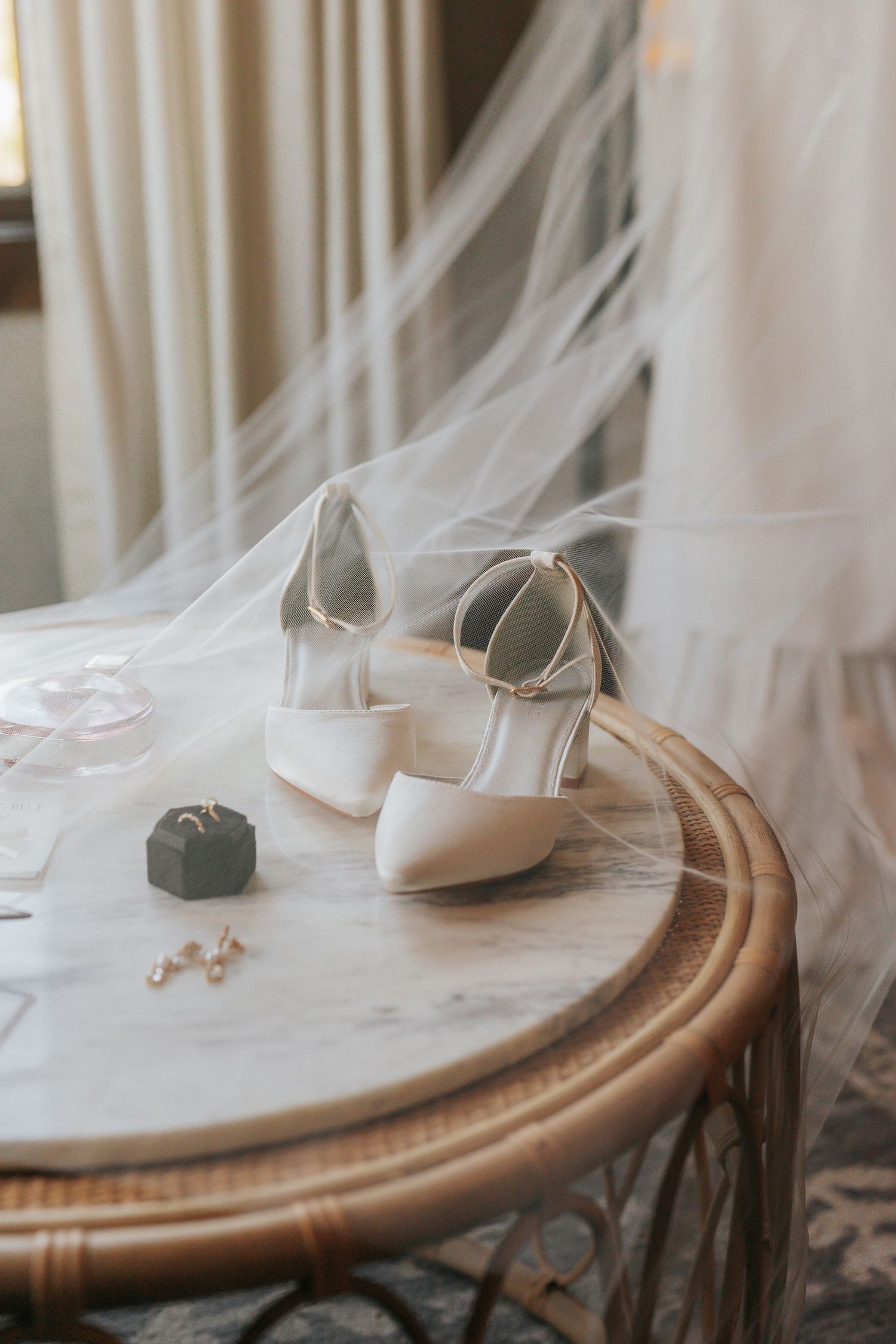White bridal heels and ring box on a marble table draped with a wedding veil.