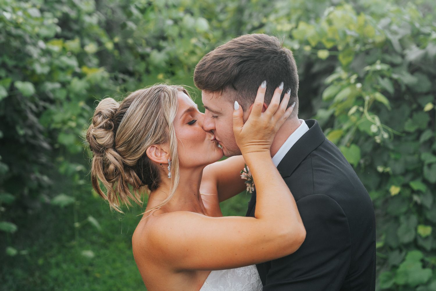 Bride and groom kissing romantically surrounded by lush green foliage during outdoor wedding portraits.