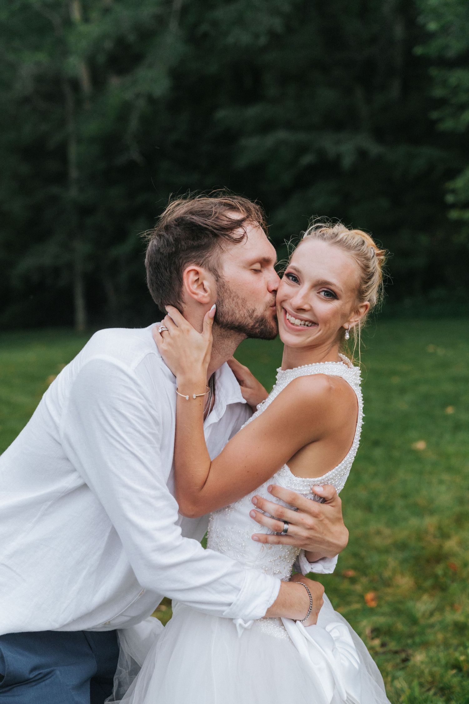Groom kisses blonde bride on cheek as she smiles during outdoor wedding portrait session.
