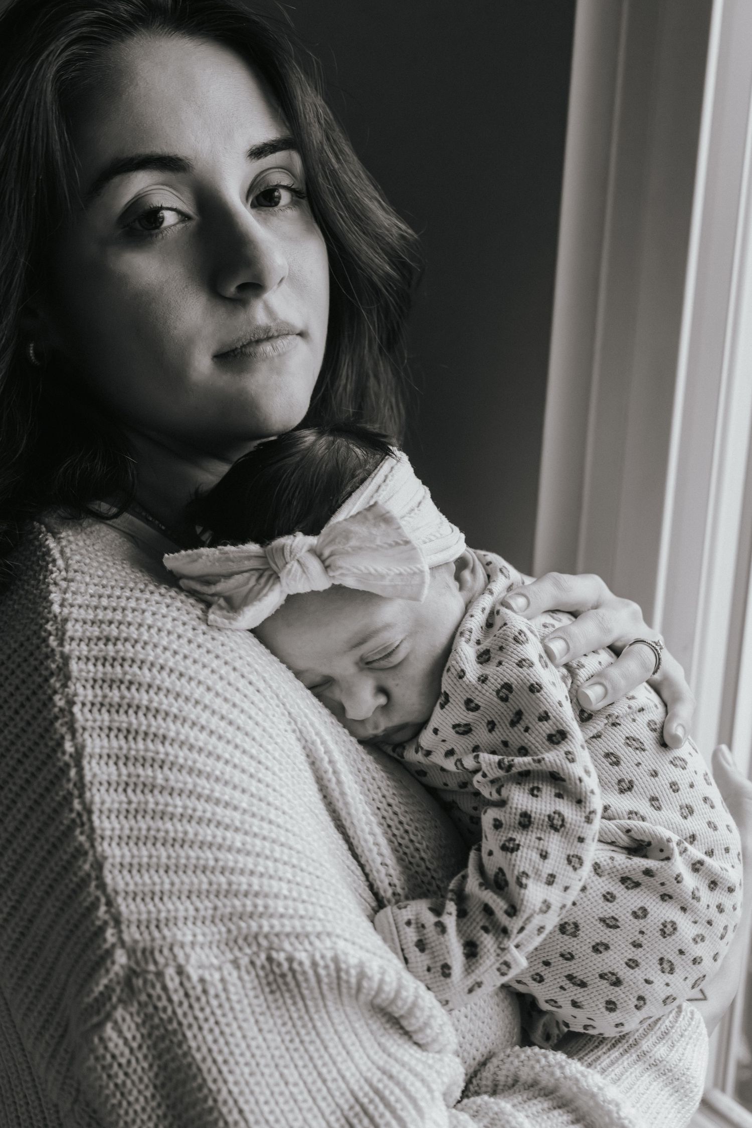 Black and white portrait of a mother tenderly holding her sleeping newborn baby near a window.
