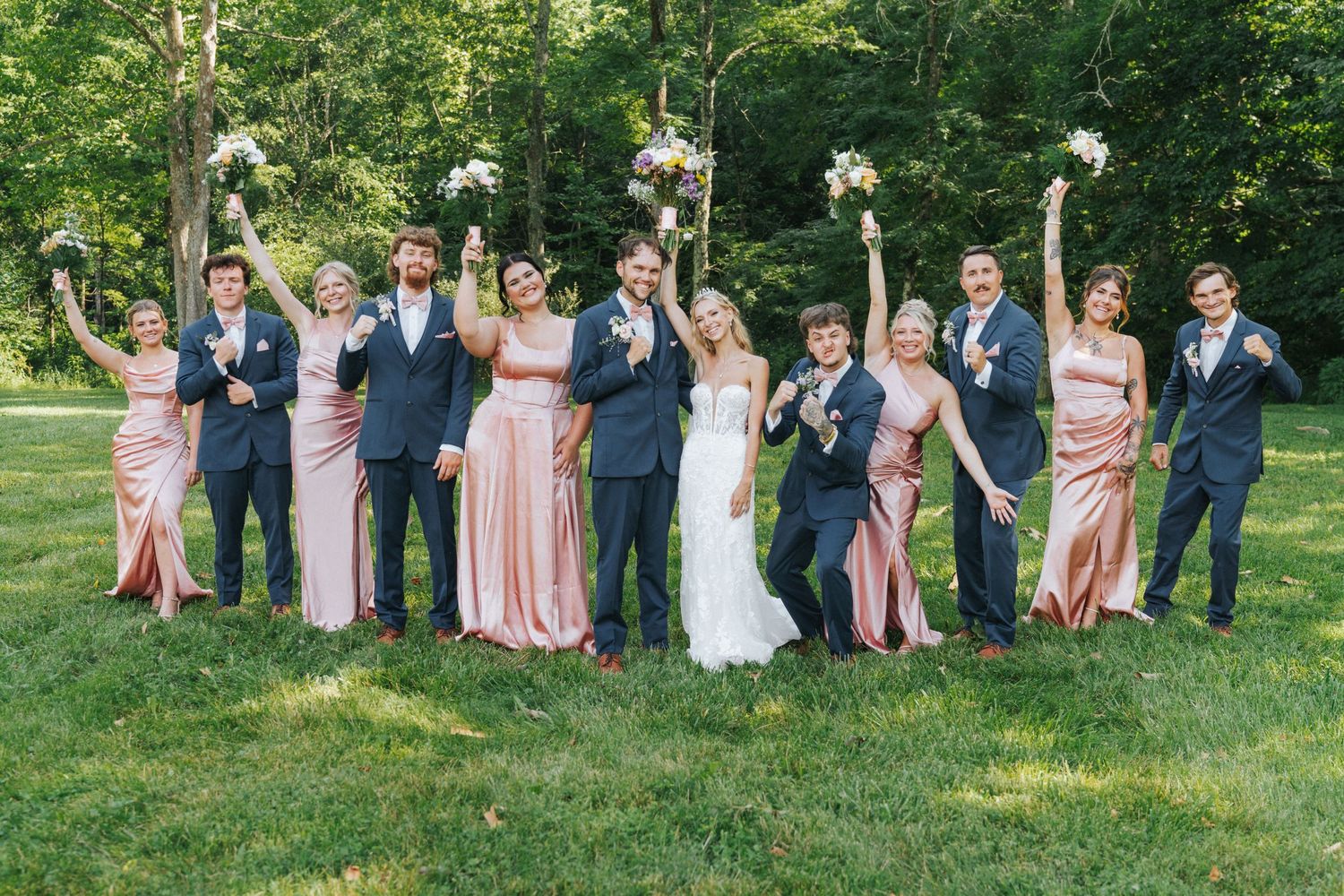 Wedding party in navy suits and blush gowns celebrating outdoors, raising bouquets with bride and groom centered on green lawn.