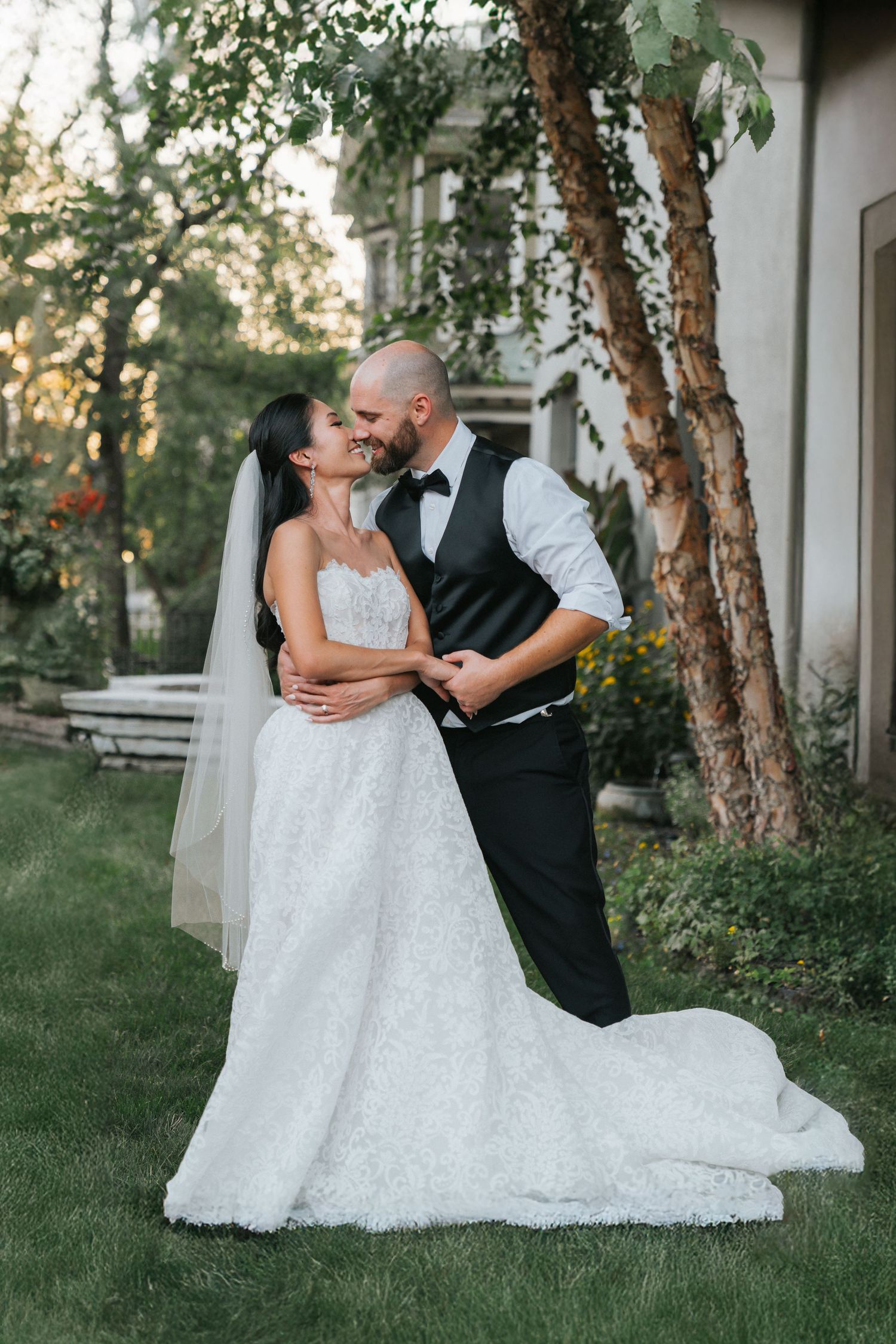 Bride in lace A-line gown and groom in black tuxedo share a kiss on green lawn beside elegant estate.