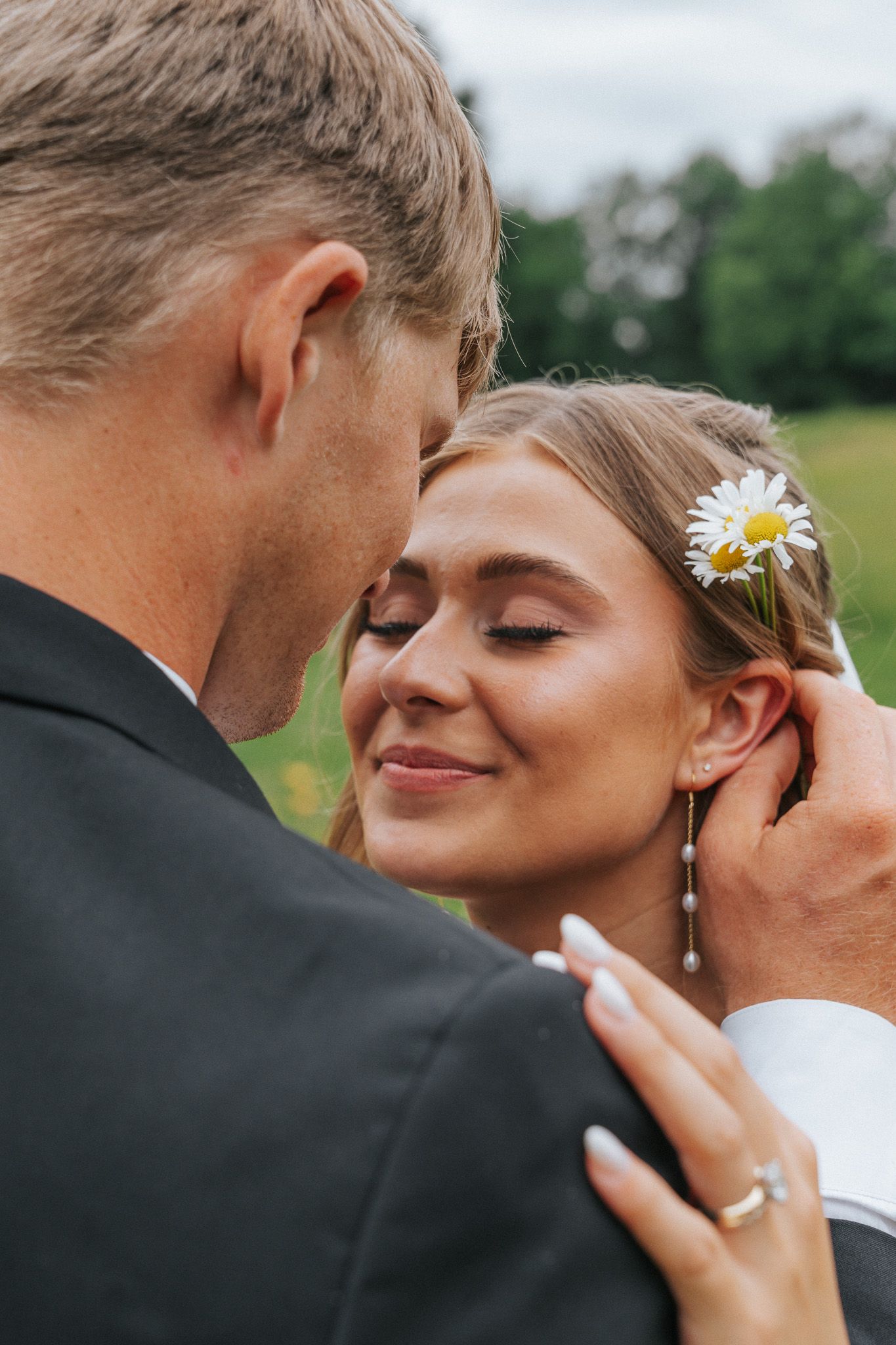 Bride and groom share a tender moment outdoors, bride wearing daisy hair accessory and pearl earrings.