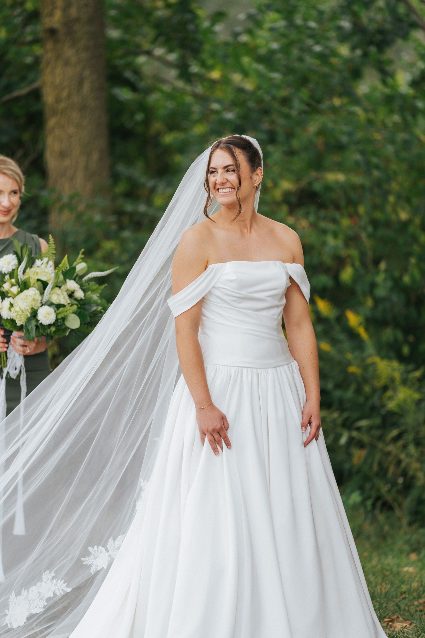 Smiling bride in off-shoulder white gown and flowing veil stands outdoors, with bridesmaid holding bouquet behind her.
