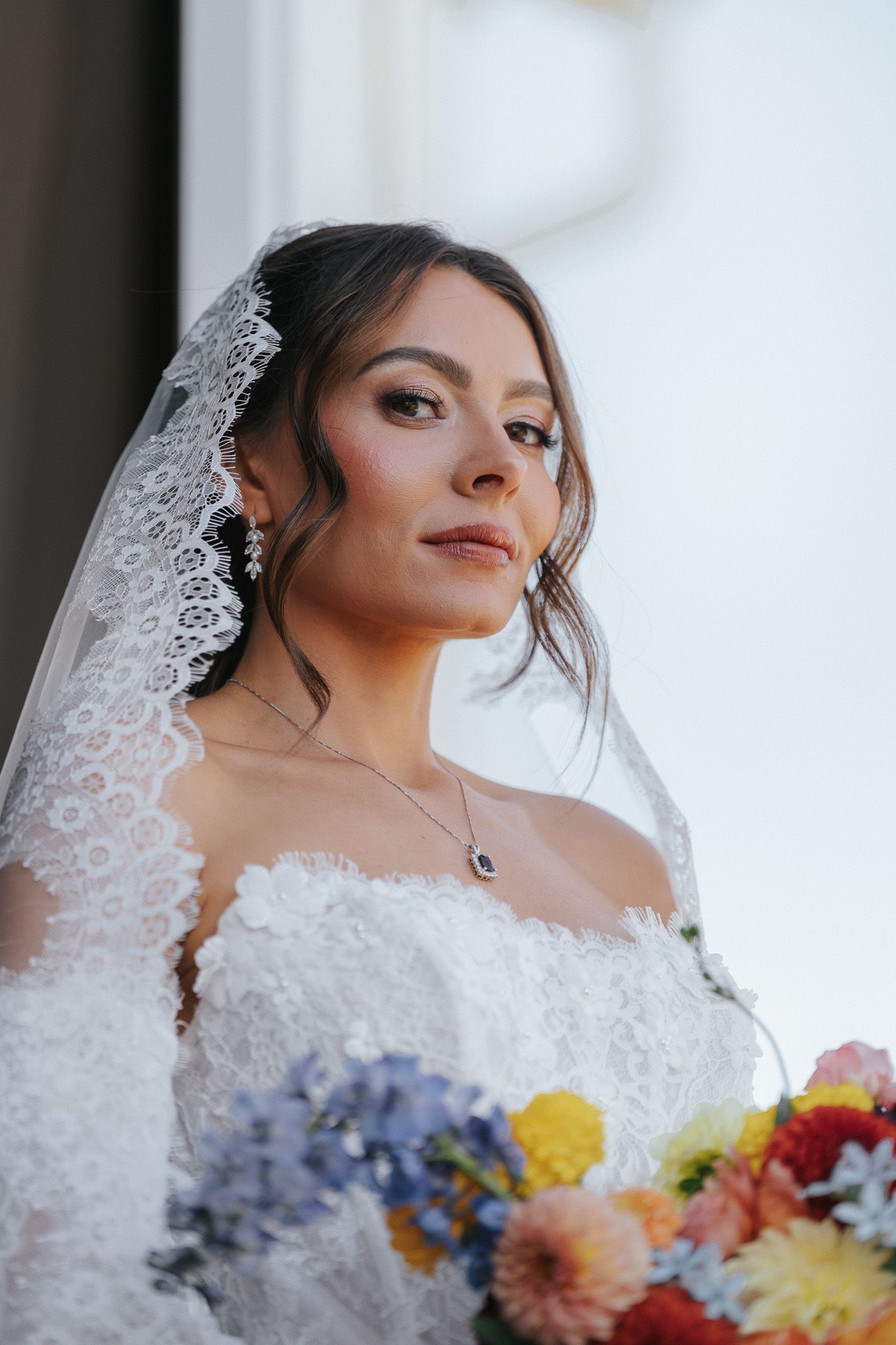 A beautiful bride in a white lace gown and veil holds a colorful bouquet, wearing elegant jewelry on her wedding day.