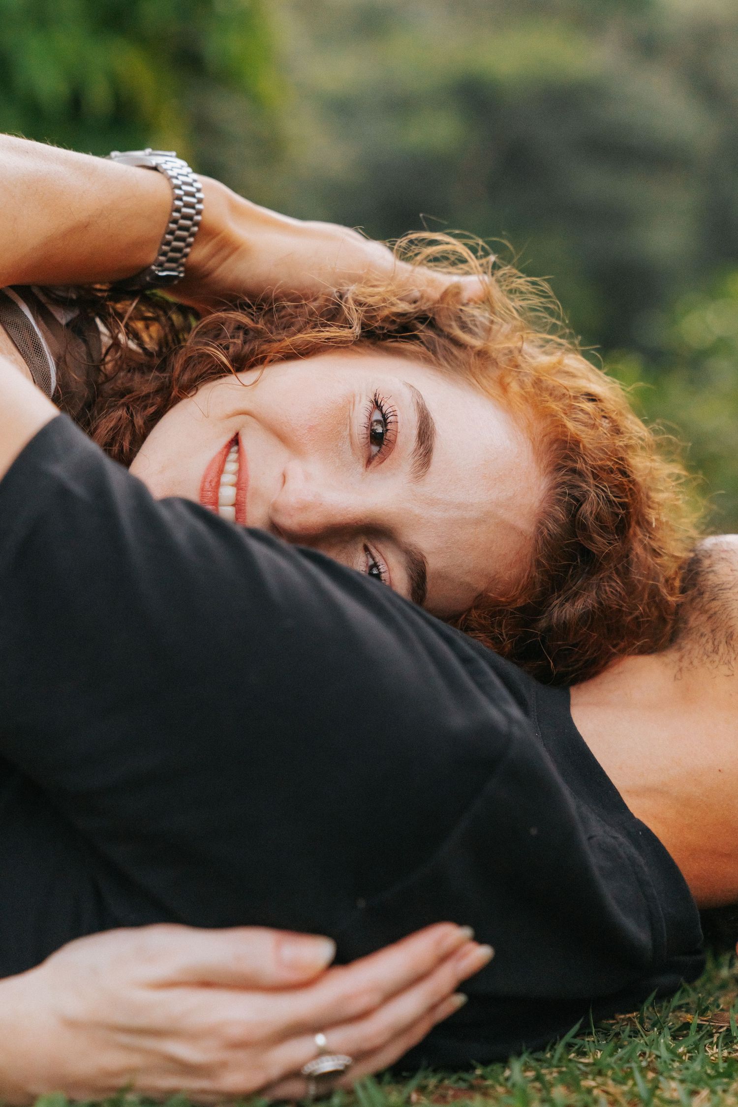 Smiling redhead woman with curly hair resting her head on her partners lap outdoors in nature.