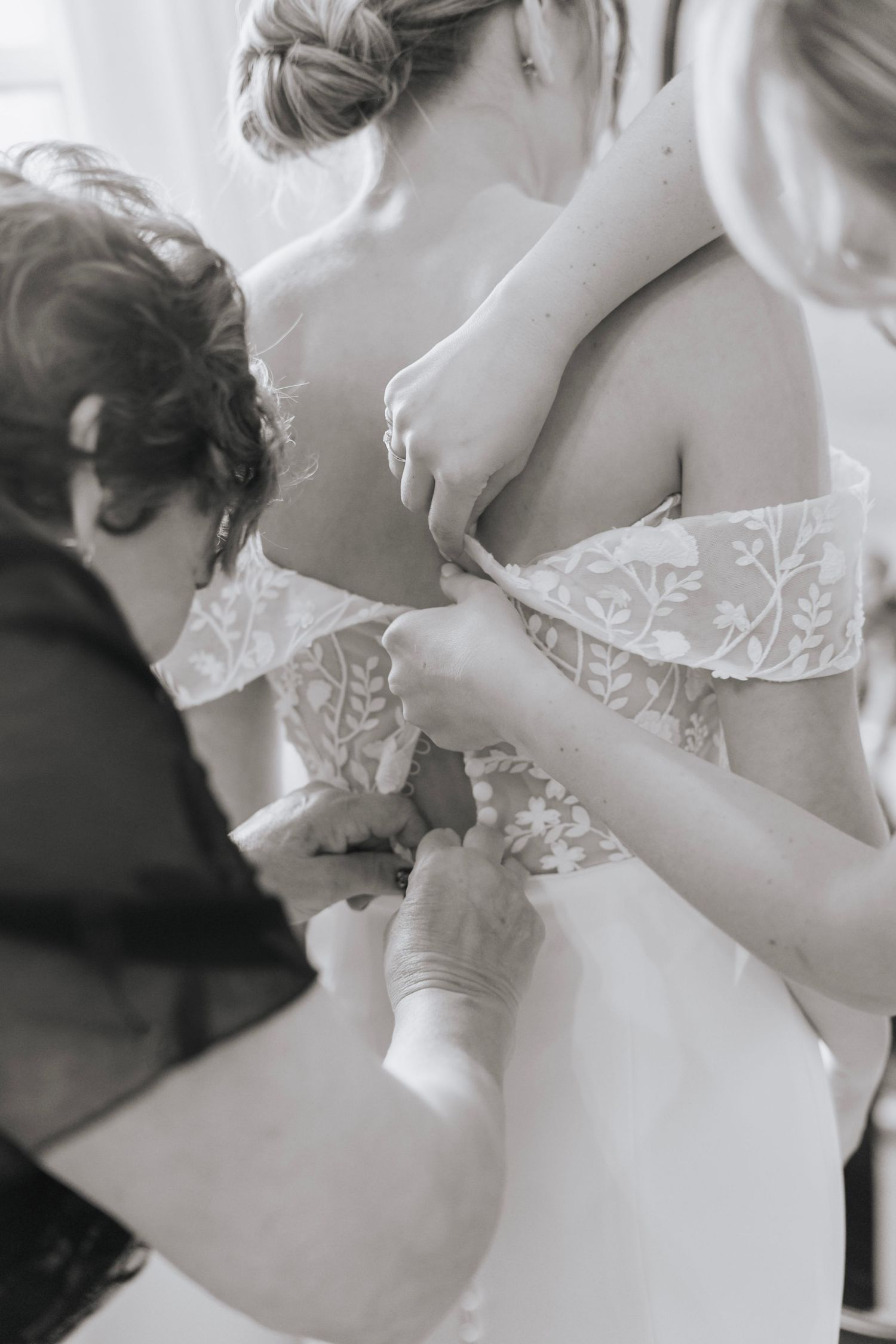 Black and white photo of bridesmaids helping bride button up her lace off-shoulder wedding dress.