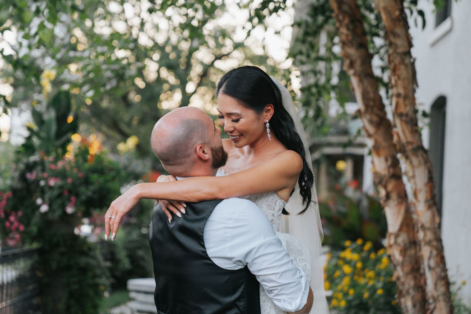 Groom lifts smiling bride in an embrace outdoors surrounded by trees and flowers.