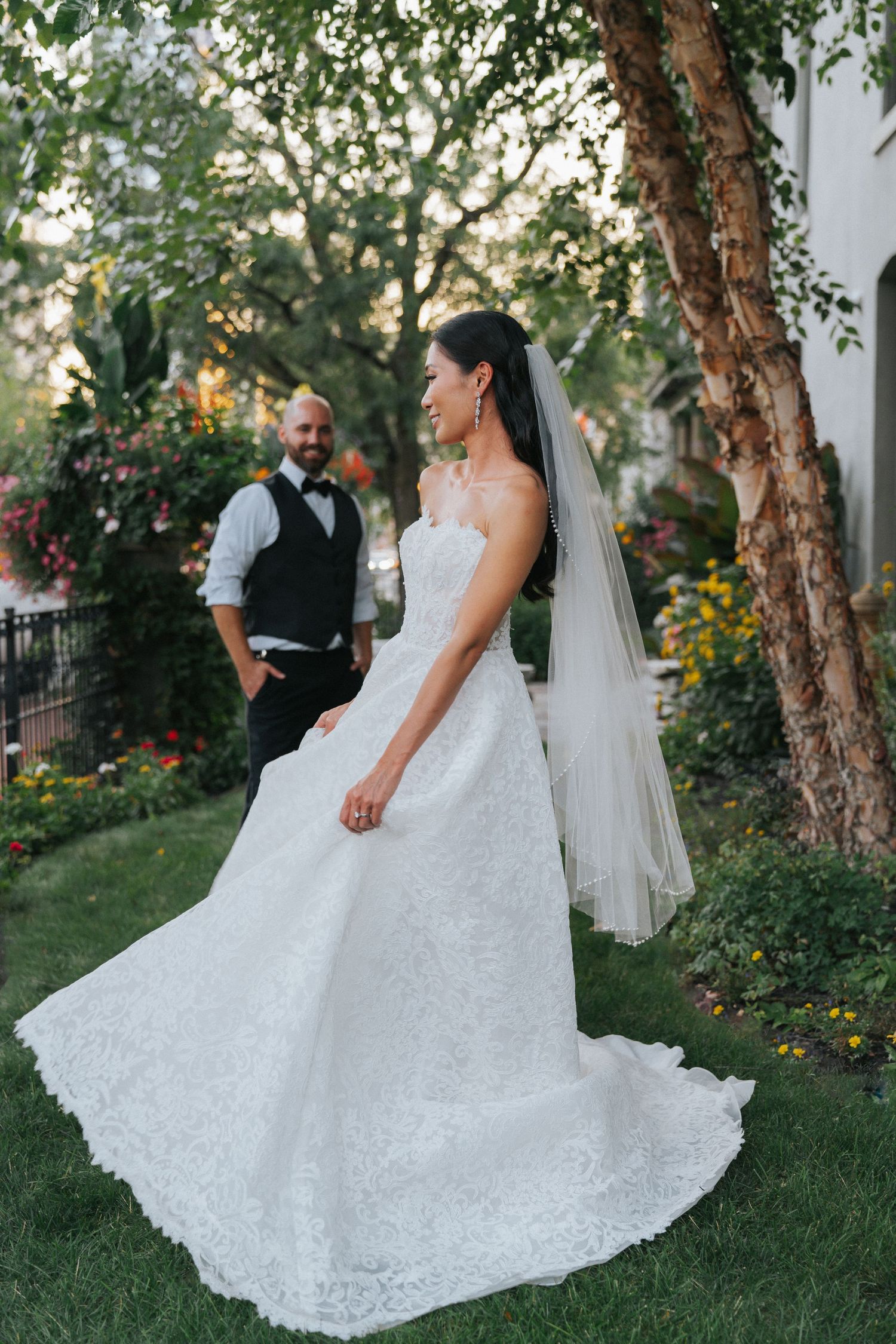 Bride in white lace gown and veil twirls in garden as groom in vest watches adoringly during outdoor wedding.