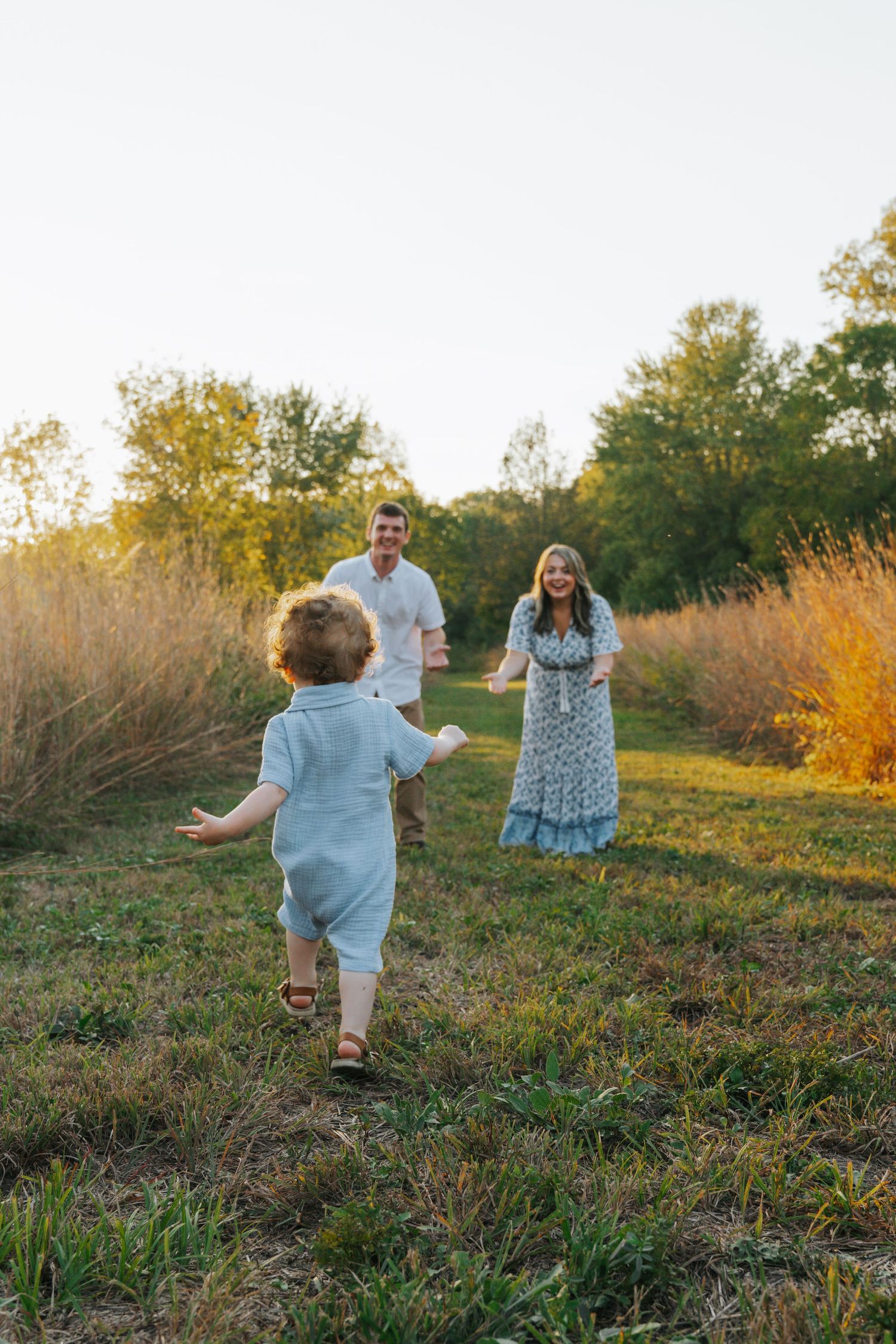 Toddler in blue romper running toward smiling parents in a sunlit autumn field at golden hour.