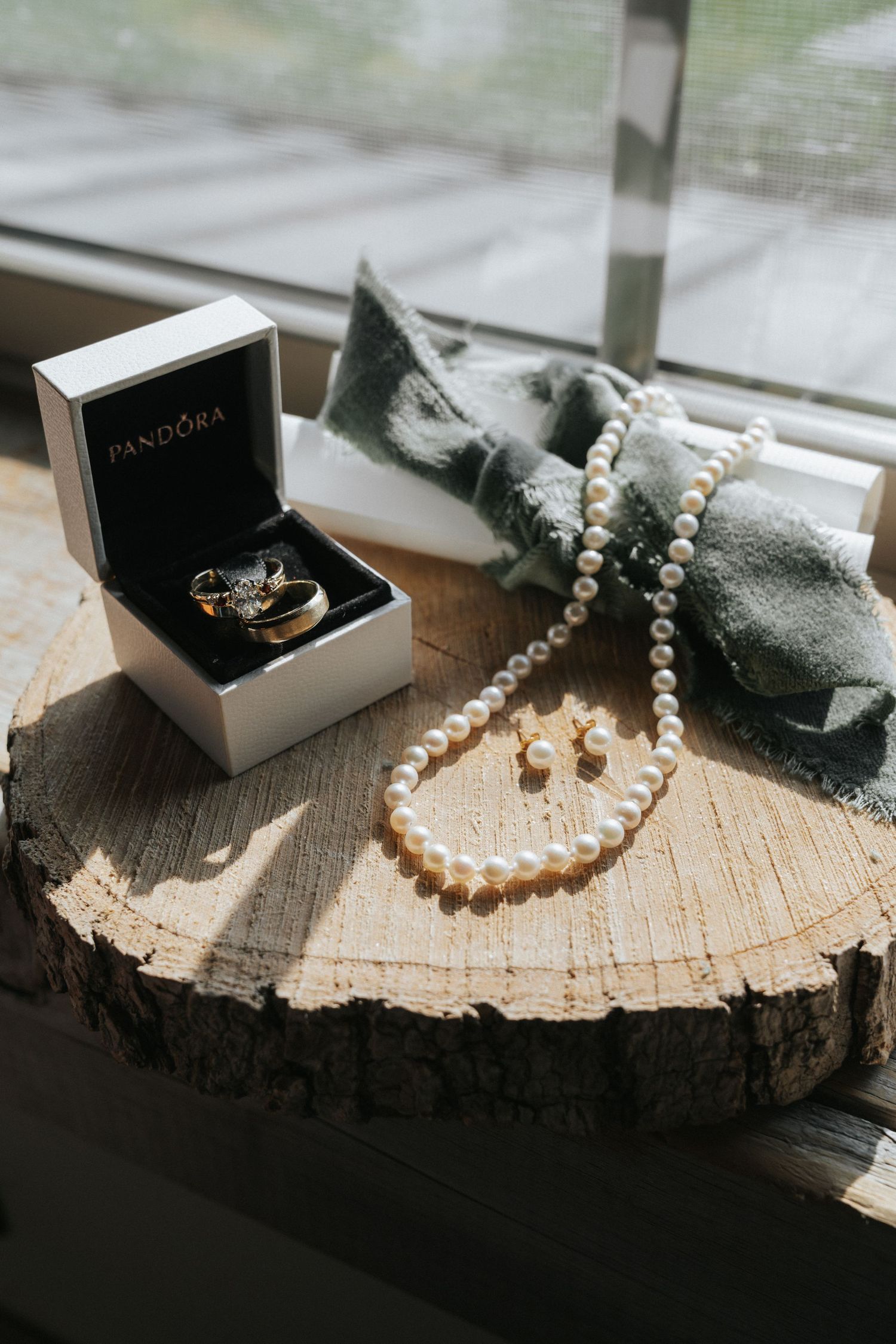 Pandora ring box with wedding bands, pearl necklace and earrings displayed on rustic wood slice.