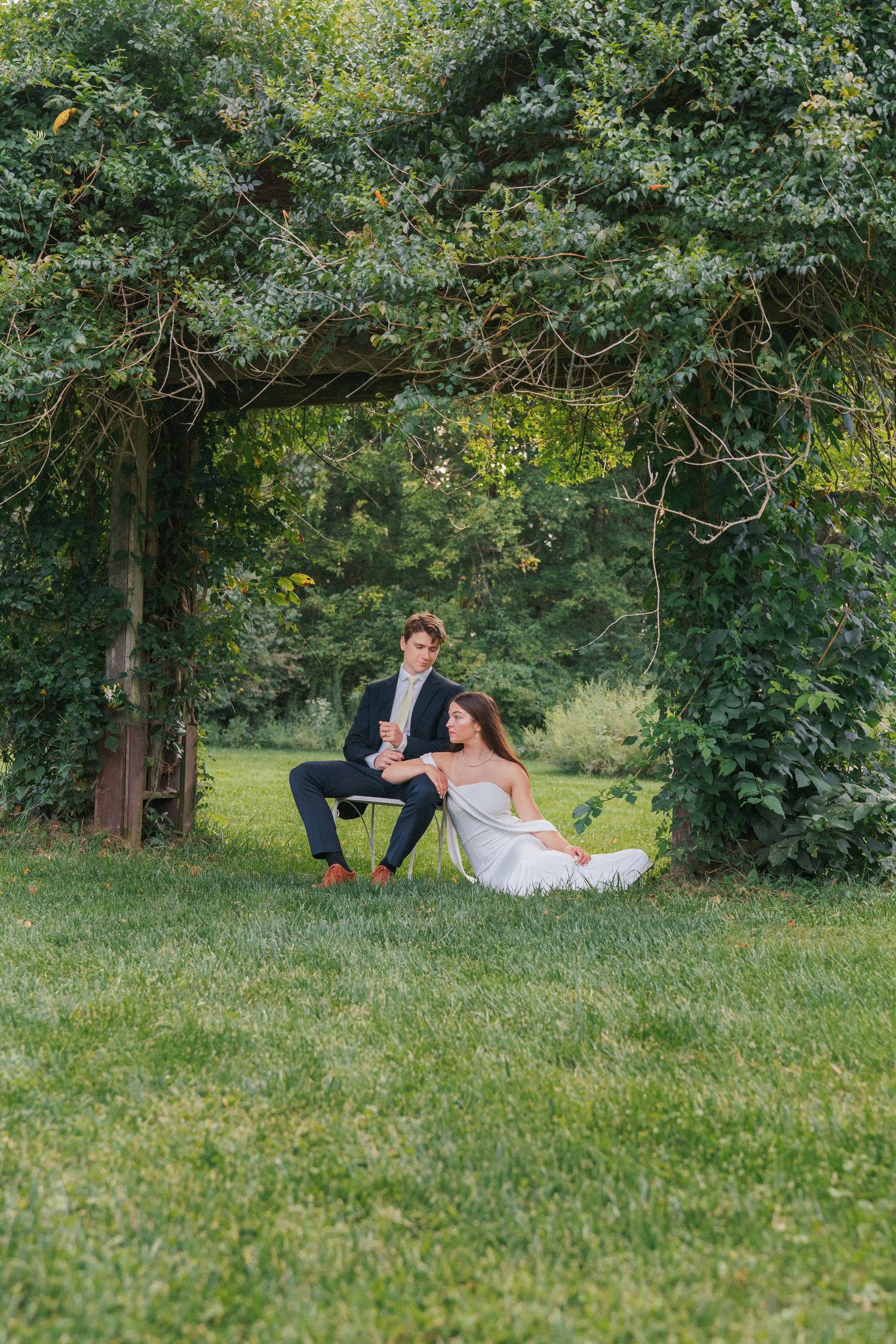 Bride in white gown sits on grass while groom in navy suit perches beside her under lush green trees.