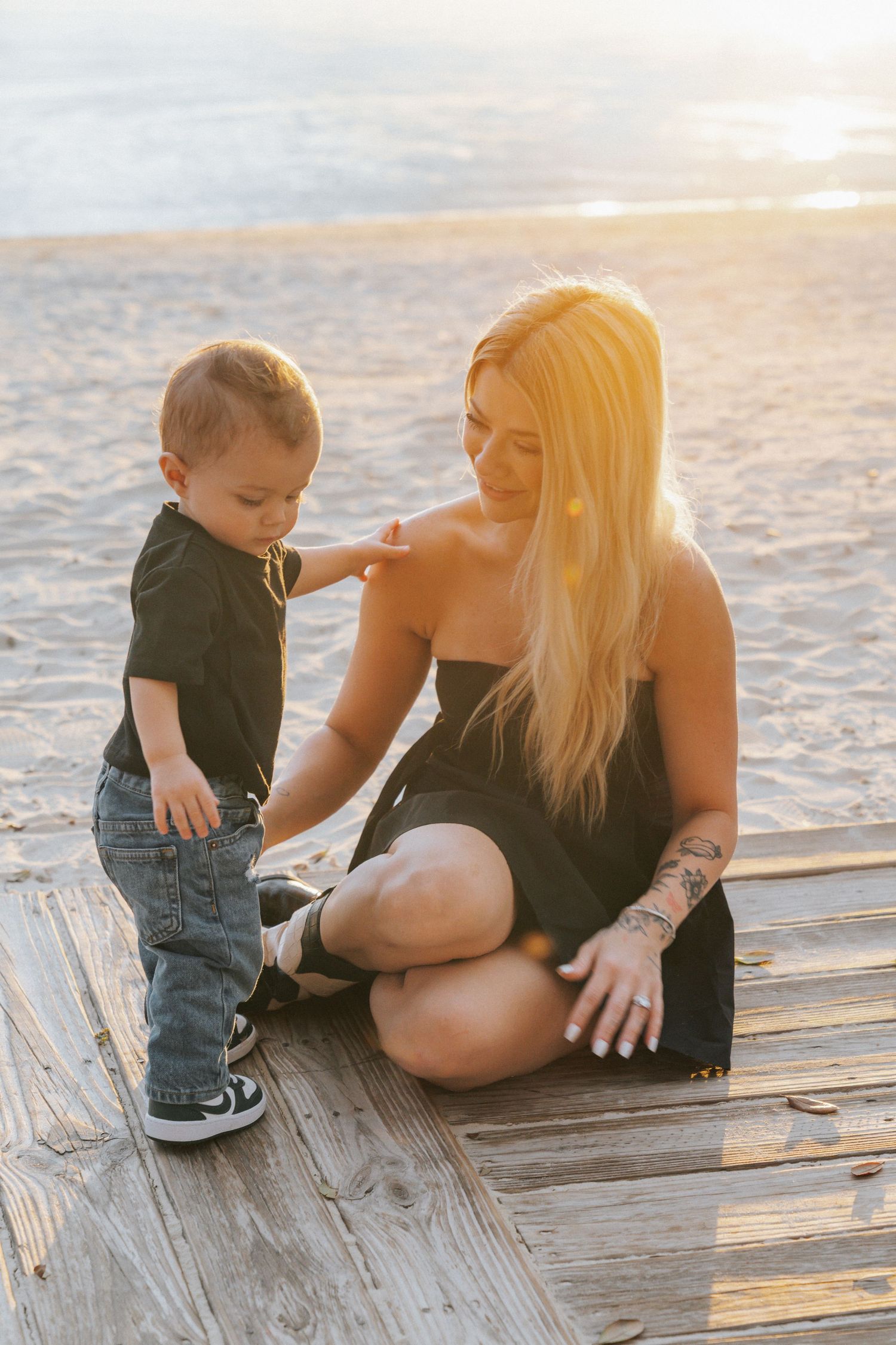 Blonde woman in black dress sits on beach boardwalk with toddler boy in jeans at golden hour sunset.