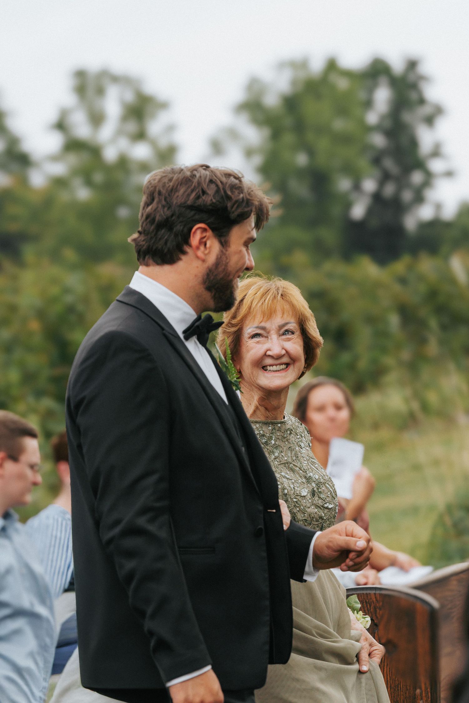 Groom in black tuxedo escorts smiling older woman at outdoor wedding ceremony with green trees in background.