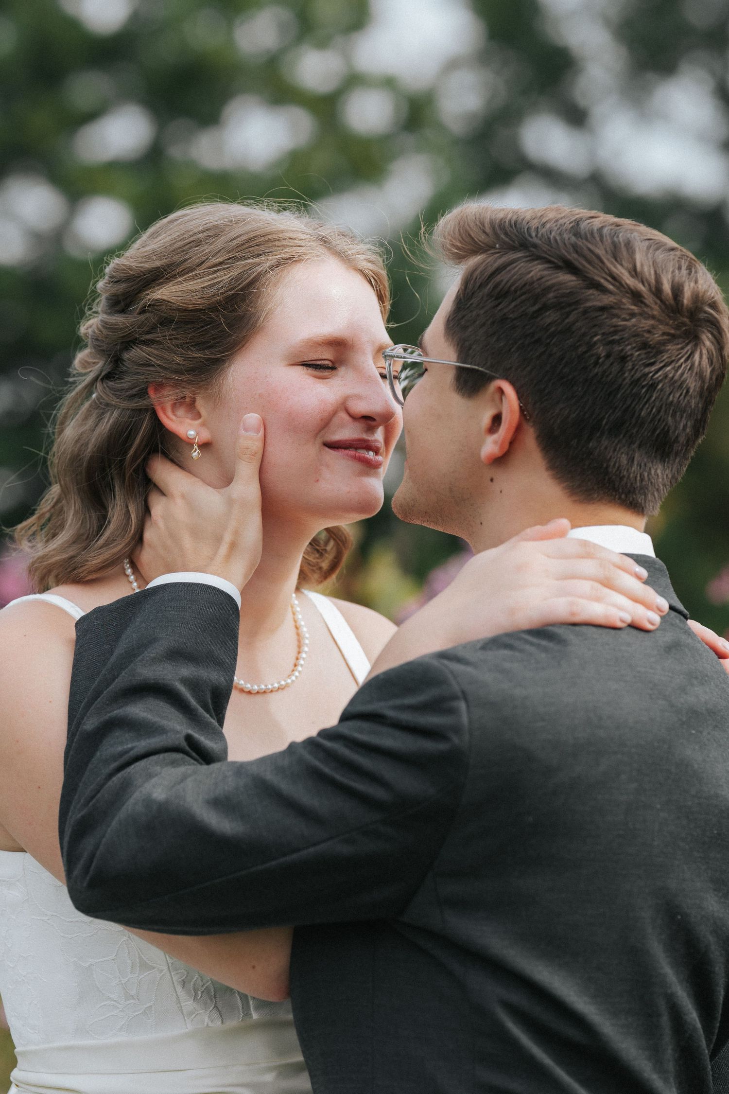 Bride and groom share a tender moment, with groom cupping bride's face as she smiles, wearing pearl necklace and white dress.