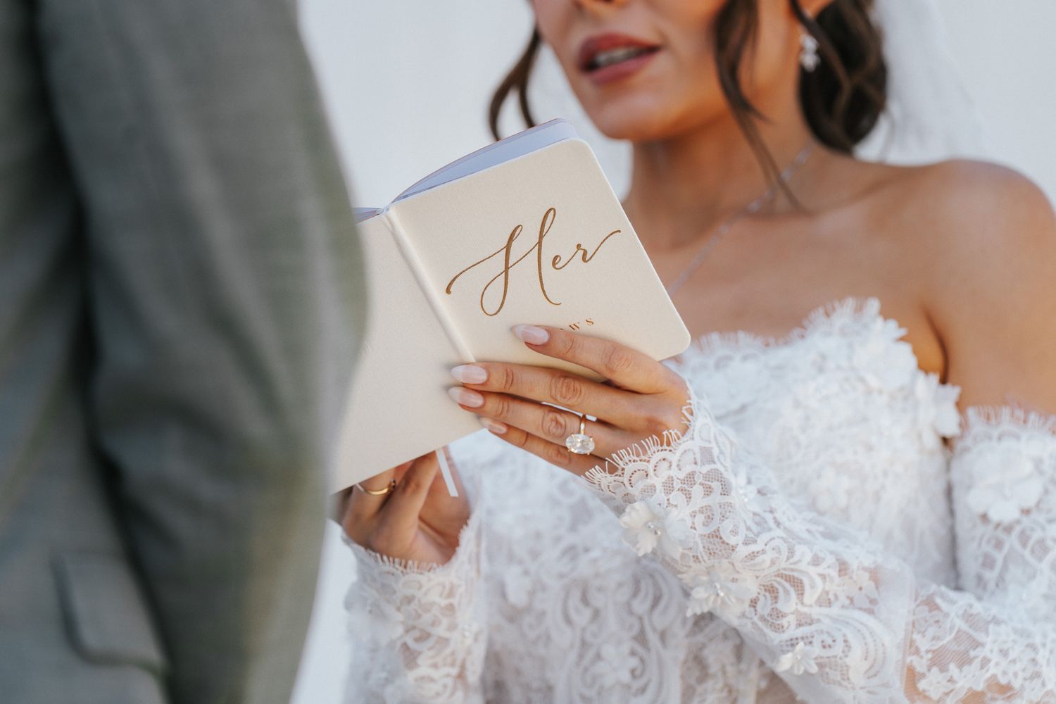 Bride in lace off-shoulder gown reads wedding vows from elegant gold booklet, showcasing diamond engagement ring.