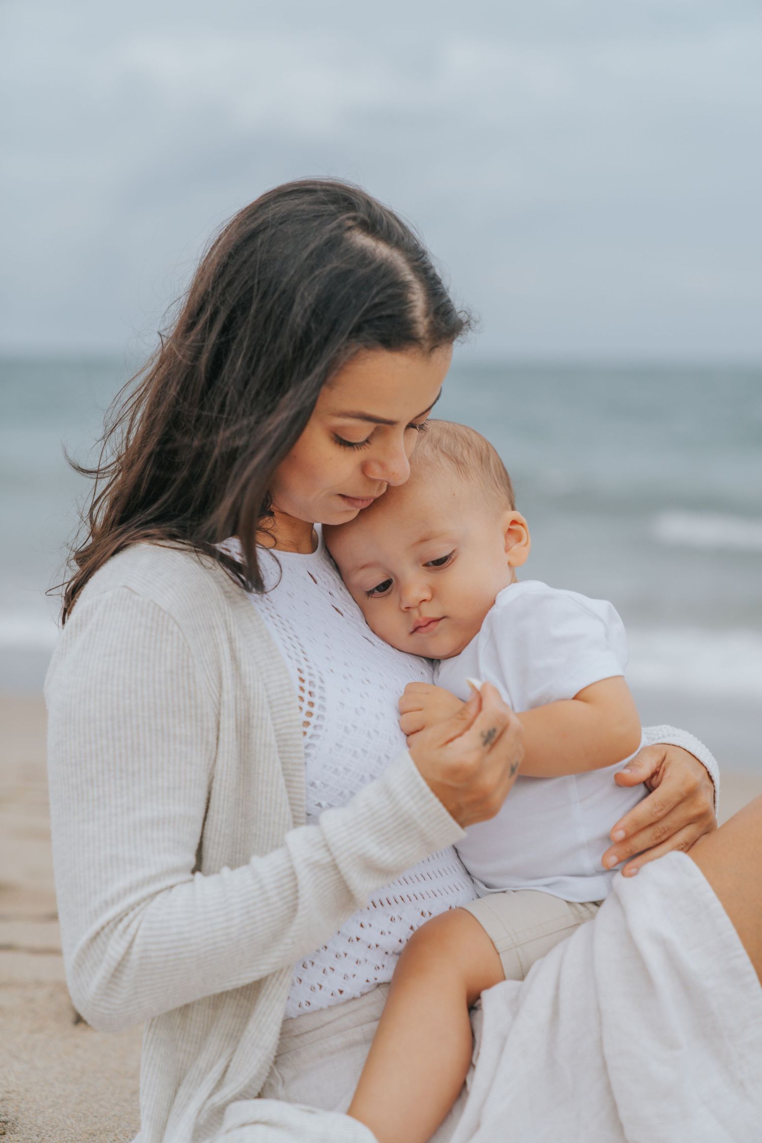Tender mother sitting on sandy beach kissing her baby while ocean waves crash in the background.