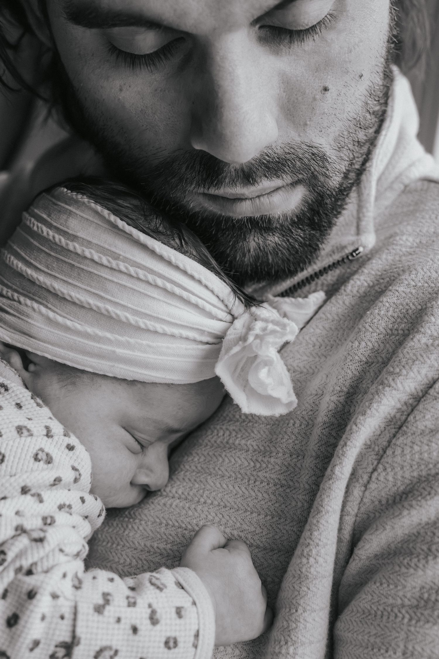 Black and white portrait of bearded father gently cradling sleeping newborn baby against his chest.