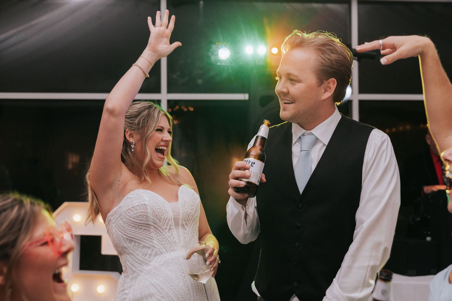 Joyful bride and groom celebrating at wedding reception, bride raising hand with excitement while groom holds champagne bottle.