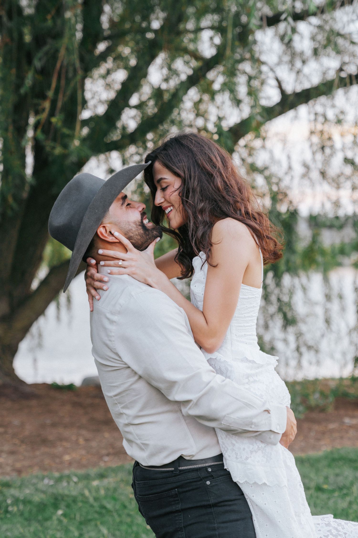 A couple shares a romantic embrace outdoors, man in gray hat lifting smiling woman in white dress near a serene lake.