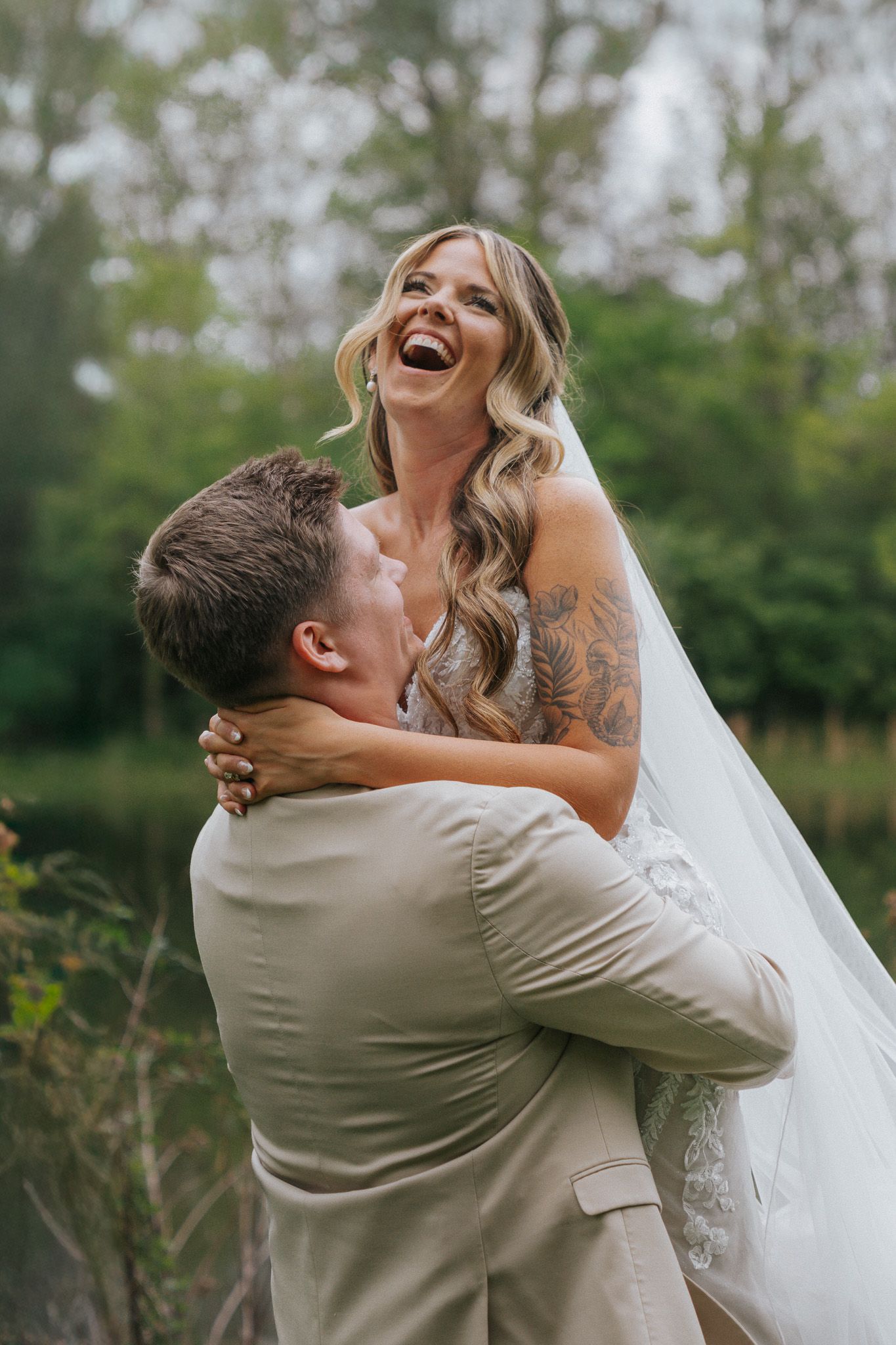 Laughing bride in veil being lifted by groom in tan suit surrounded by green trees outdoors.