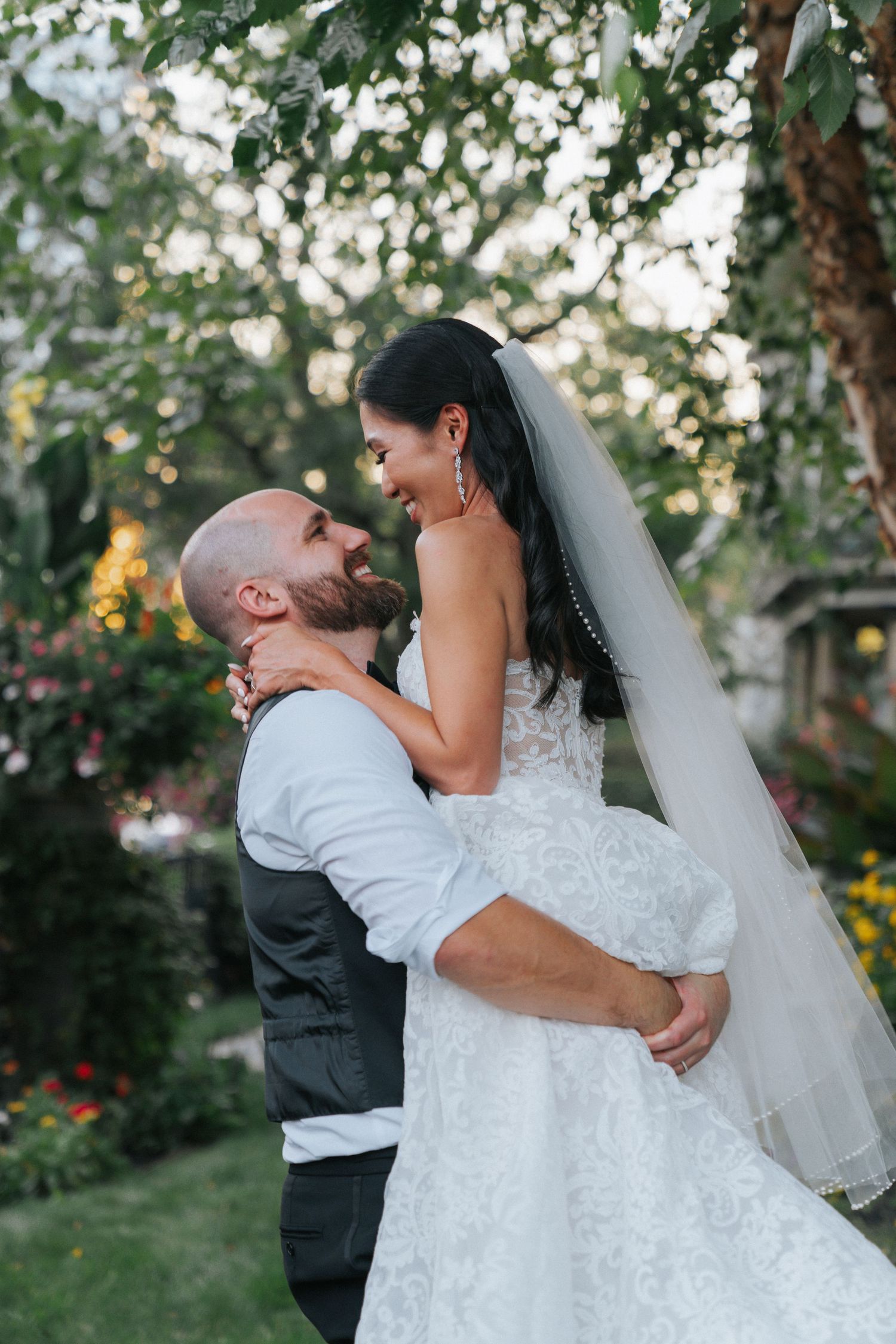 Groom lifting smiling bride in lace gown under lush garden trees at dusk.