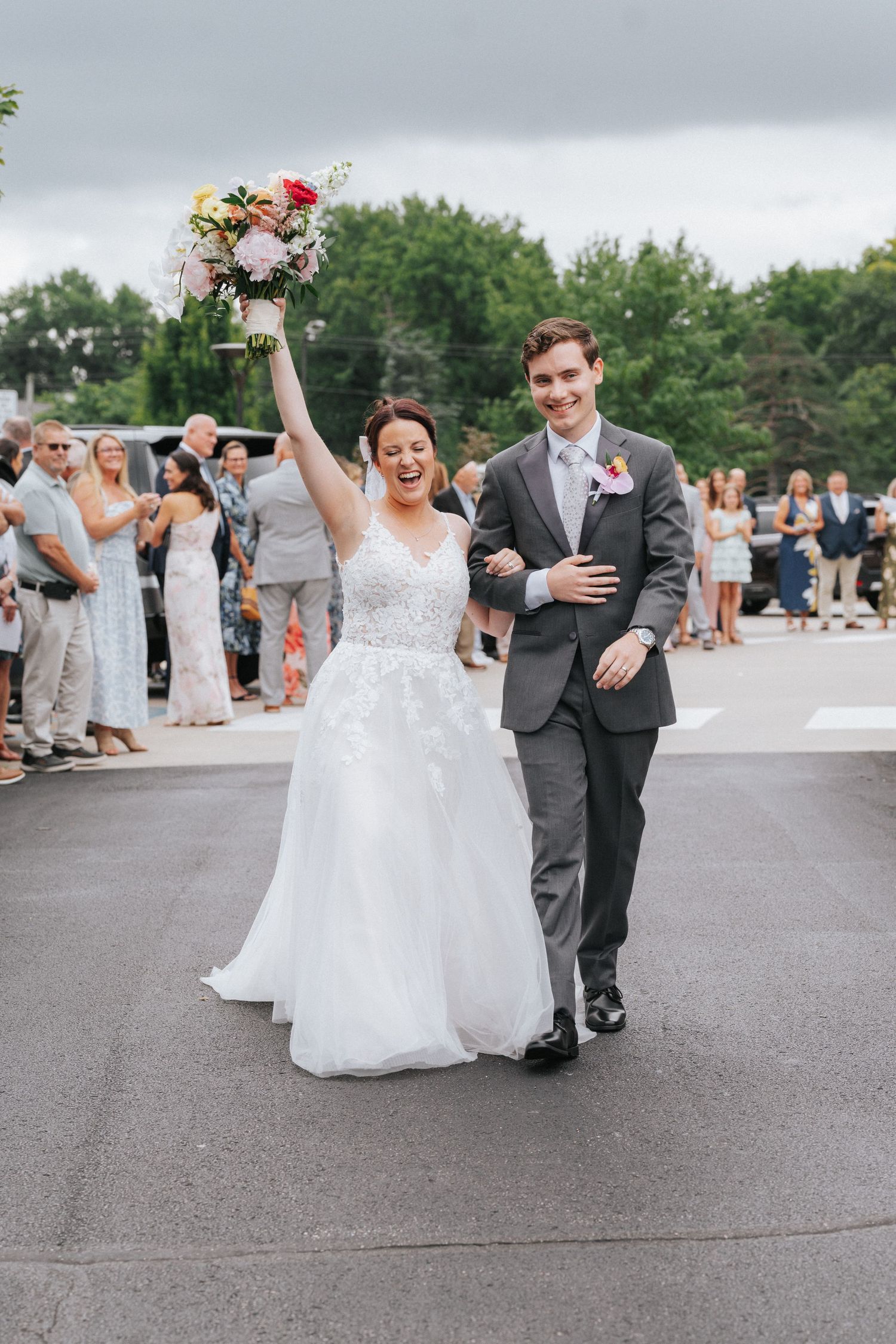 Joyful bride raising bouquet while walking with groom after wedding ceremony outdoors.
