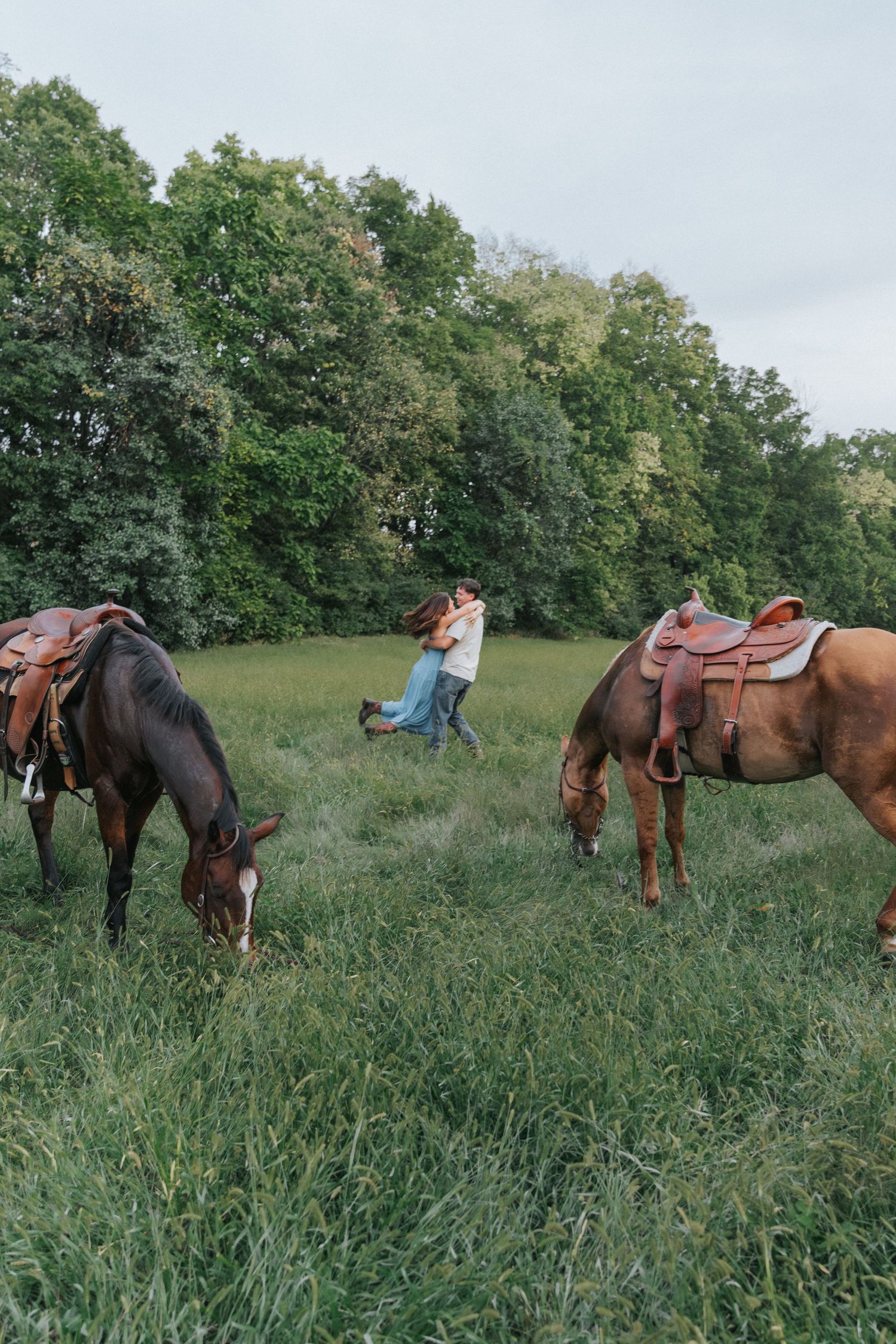 Romantic couple embracing in a green meadow with two saddled horses grazing nearby.