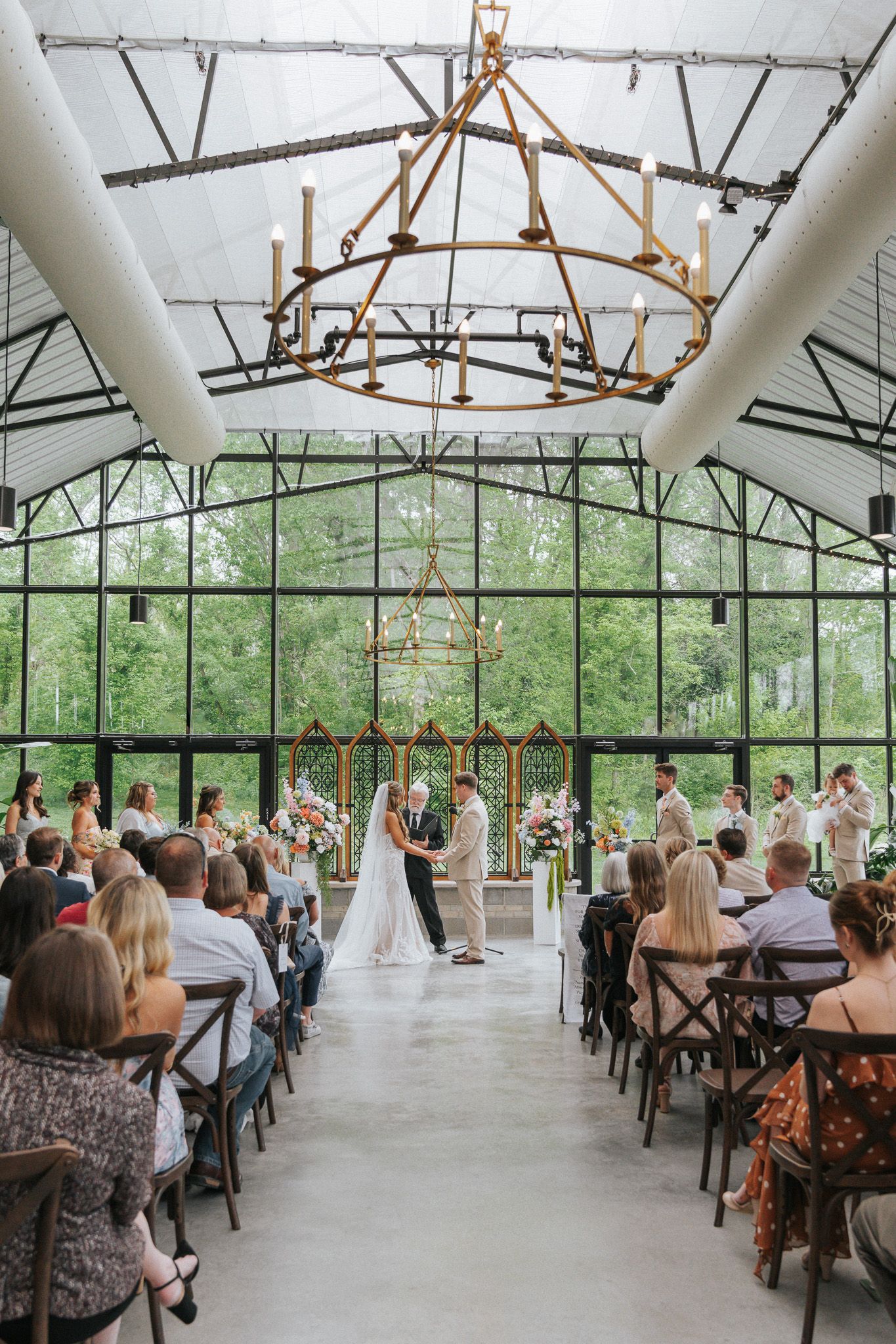 Elegant greenhouse wedding ceremony with chandelier, glass walls, and guests seated along a floral aisle.