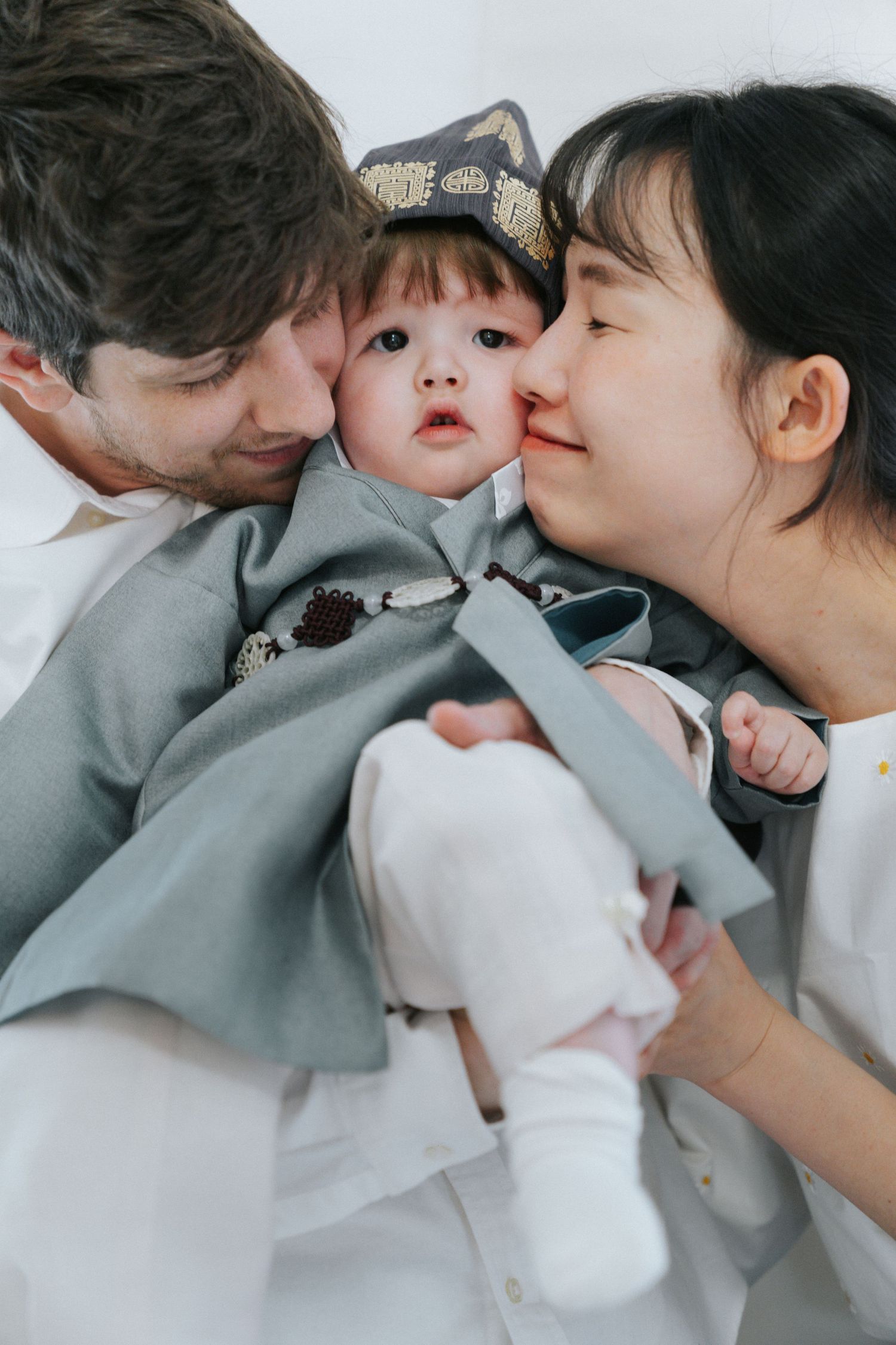 Loving interracial parents kissing their baby dressed in traditional Korean hanbok on both cheeks.