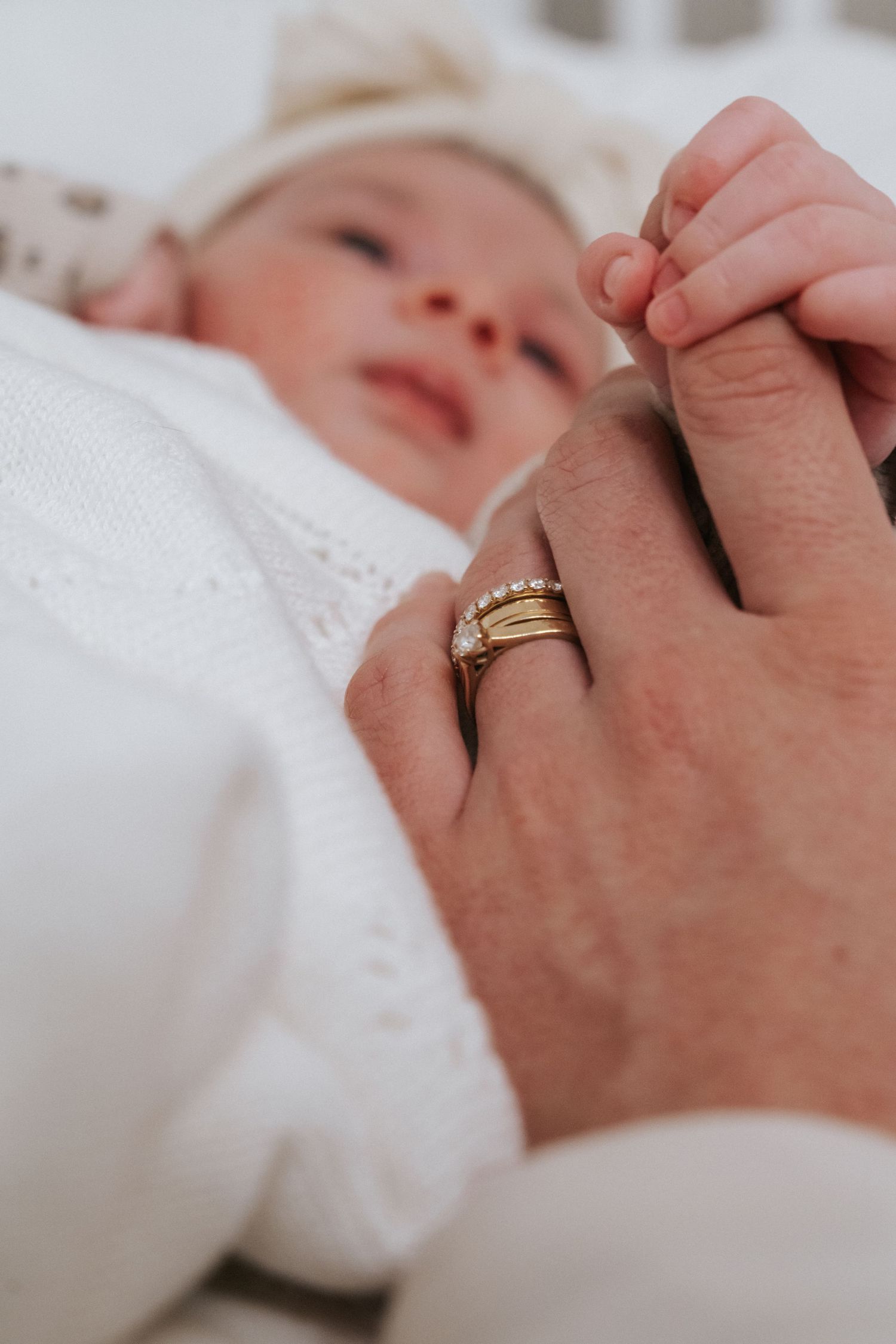 Newborn baby grasping parent's finger adorned with gold stacking rings while lying in white wrap.