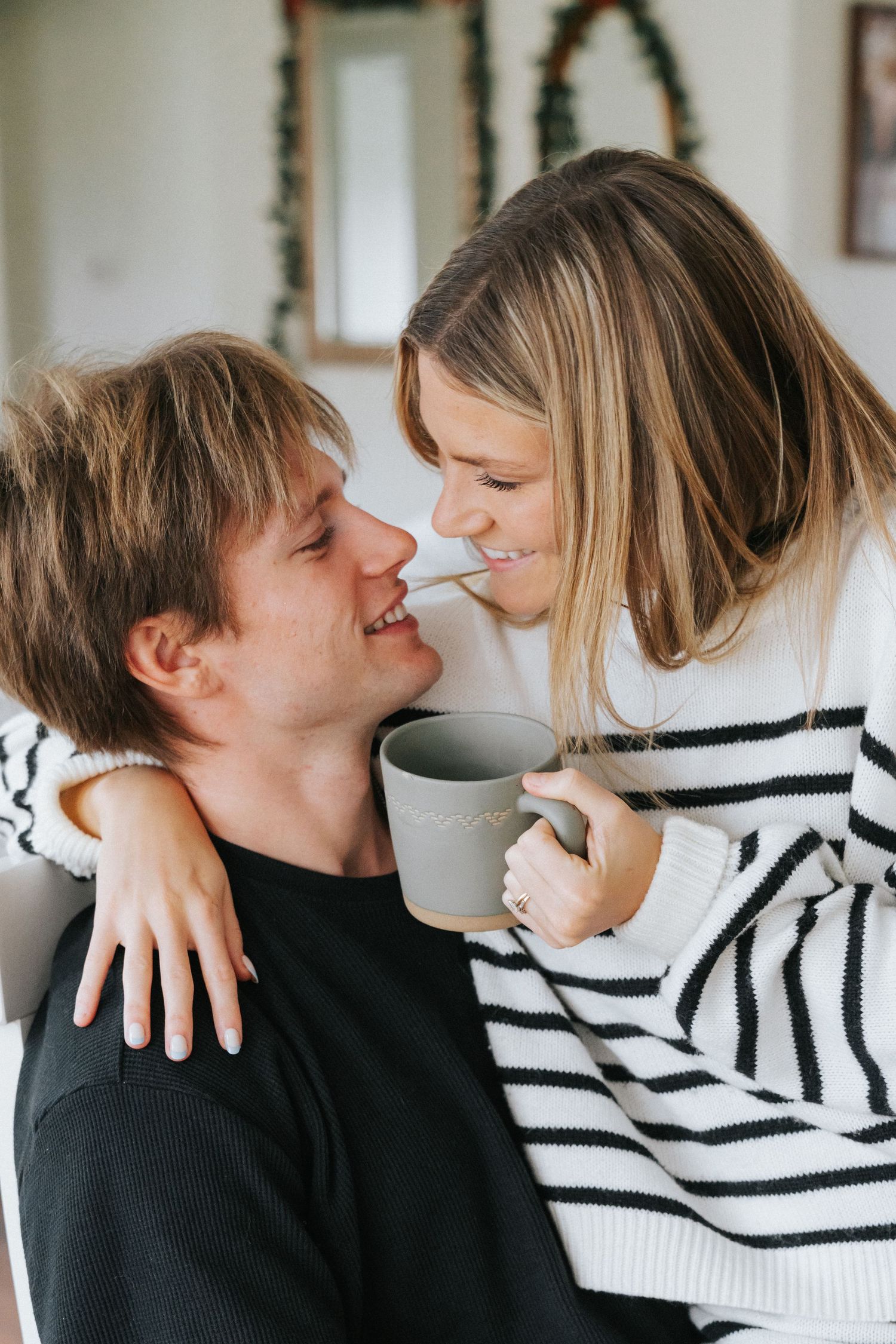 Couple smiling and gazing at each other affectionately indoors, woman holding a gray coffee mug.