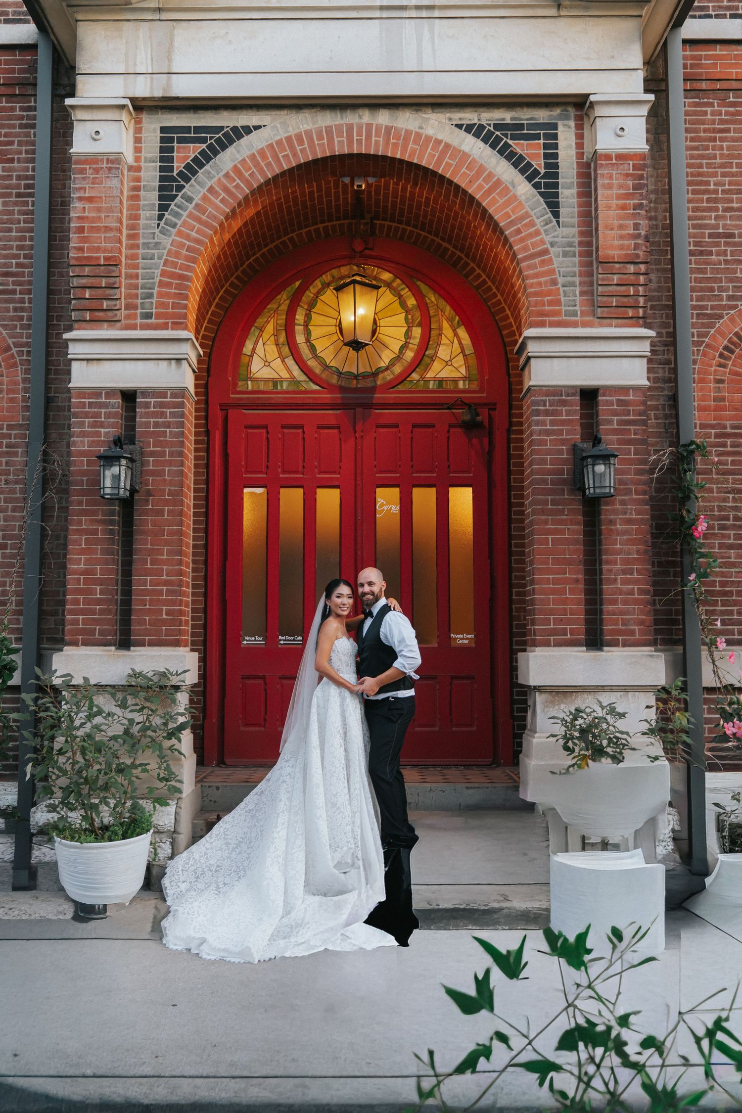 Bride and groom posing in front of elegant red doors with stained glass arch at a brick wedding venue.