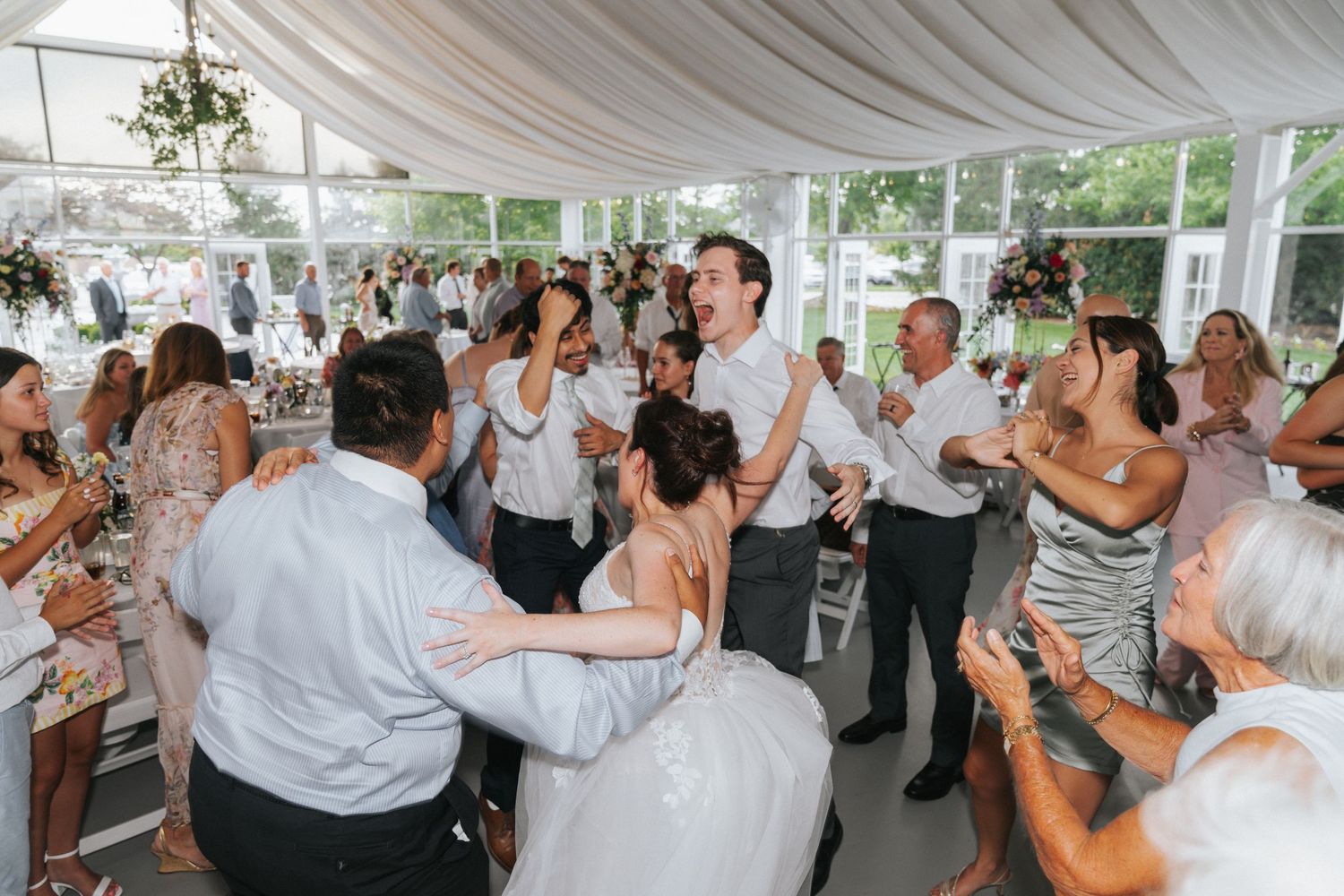 Joyful wedding reception dance floor with bride in white gown surrounded by celebrating guests in elegant marquee venue.