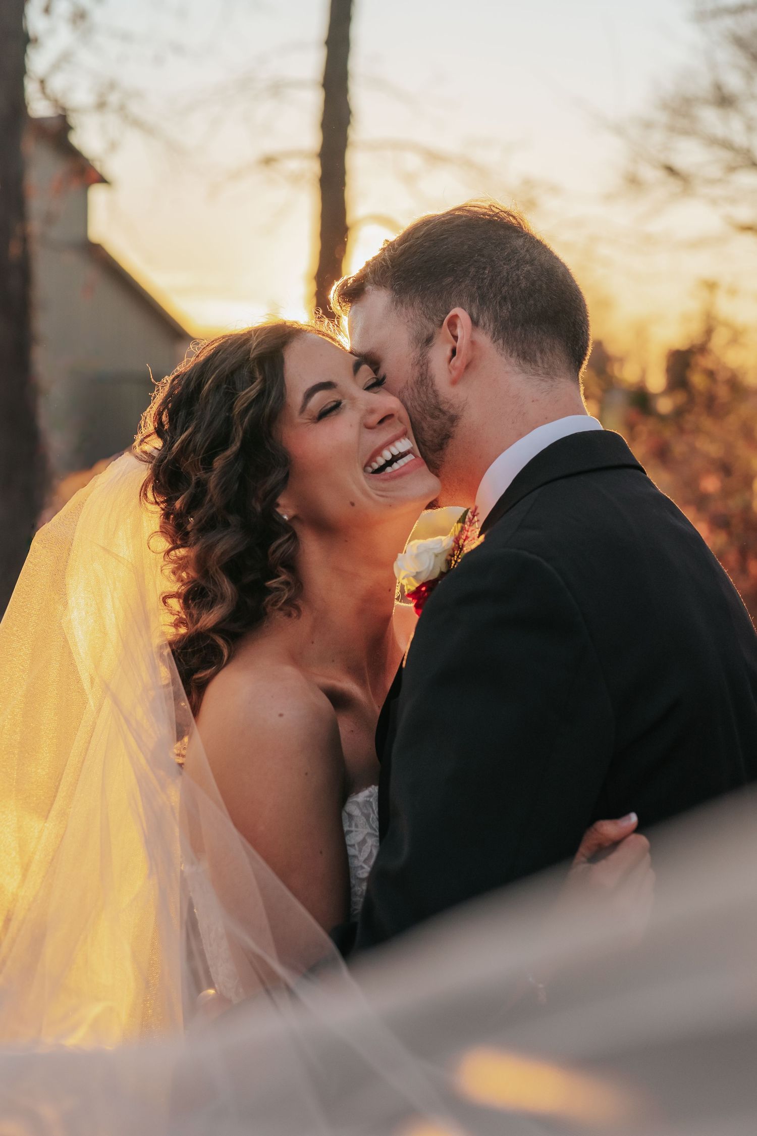 Bride laughing joyfully as groom kisses her cheek during golden sunset wedding portrait.