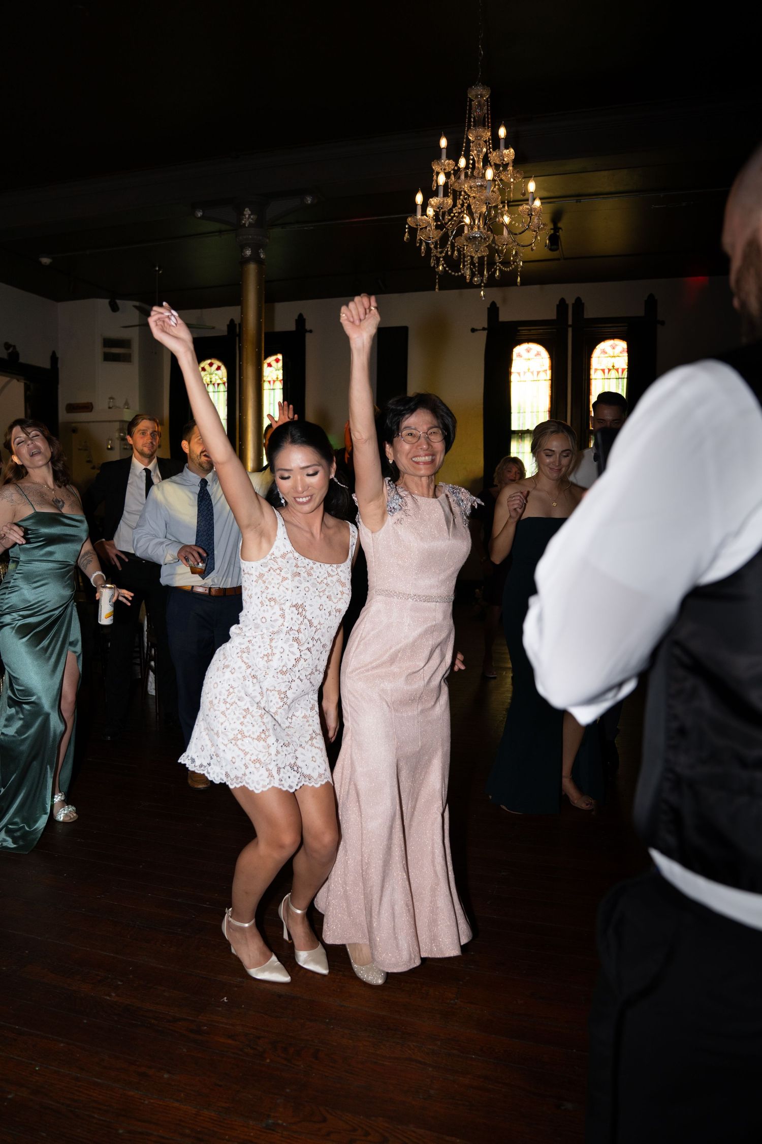 Two women in elegant dresses raise their arms joyfully on a wedding reception dance floor under a chandelier.