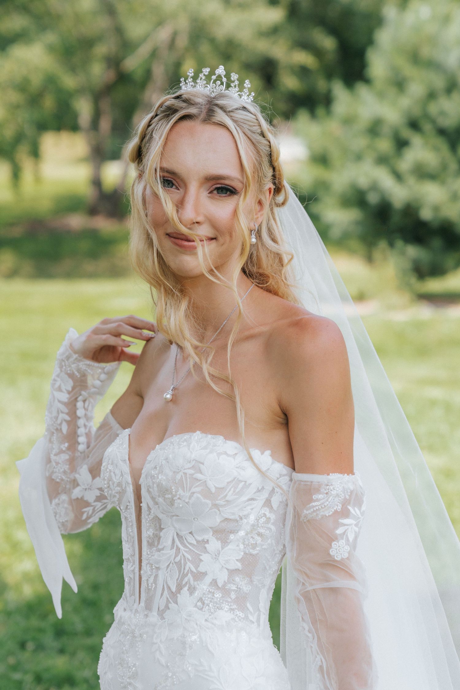 Elegant bride in lace off-shoulder gown with sheer gloves, crystal tiara, and veil posing outdoors.