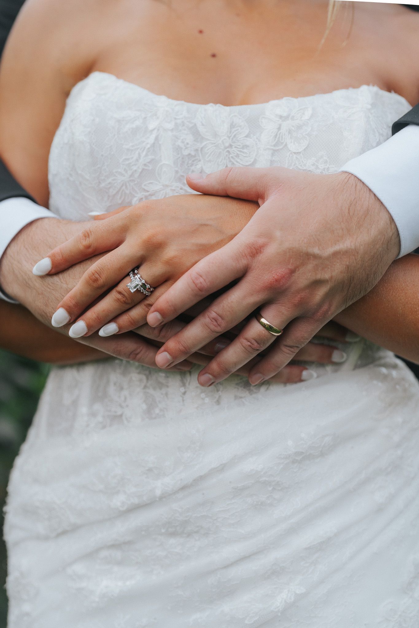 Close-up of bride and groom hands showing wedding rings, groom embracing bride in lace dress.