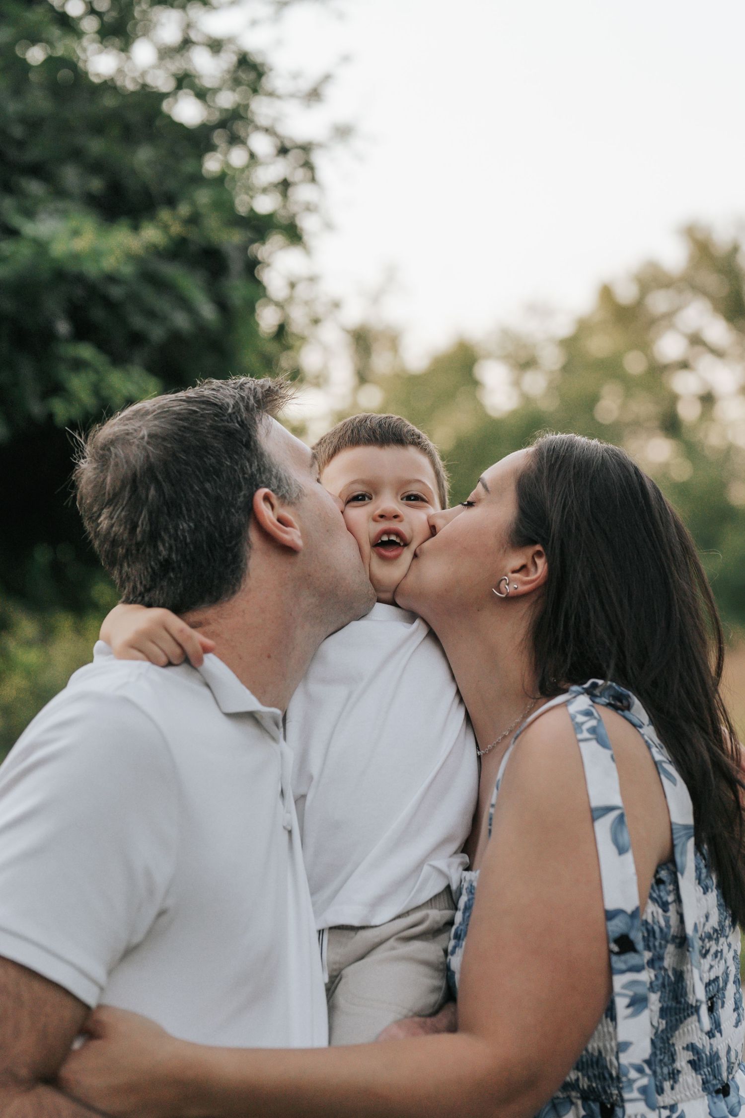 Loving parents kissing their giggling toddler son on both cheeks outdoors in a lush green park.