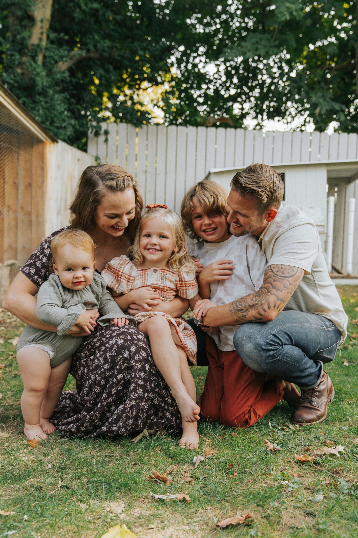 Happy family of five sitting together on lawn in backyard, parents cuddling three young children outdoors.
