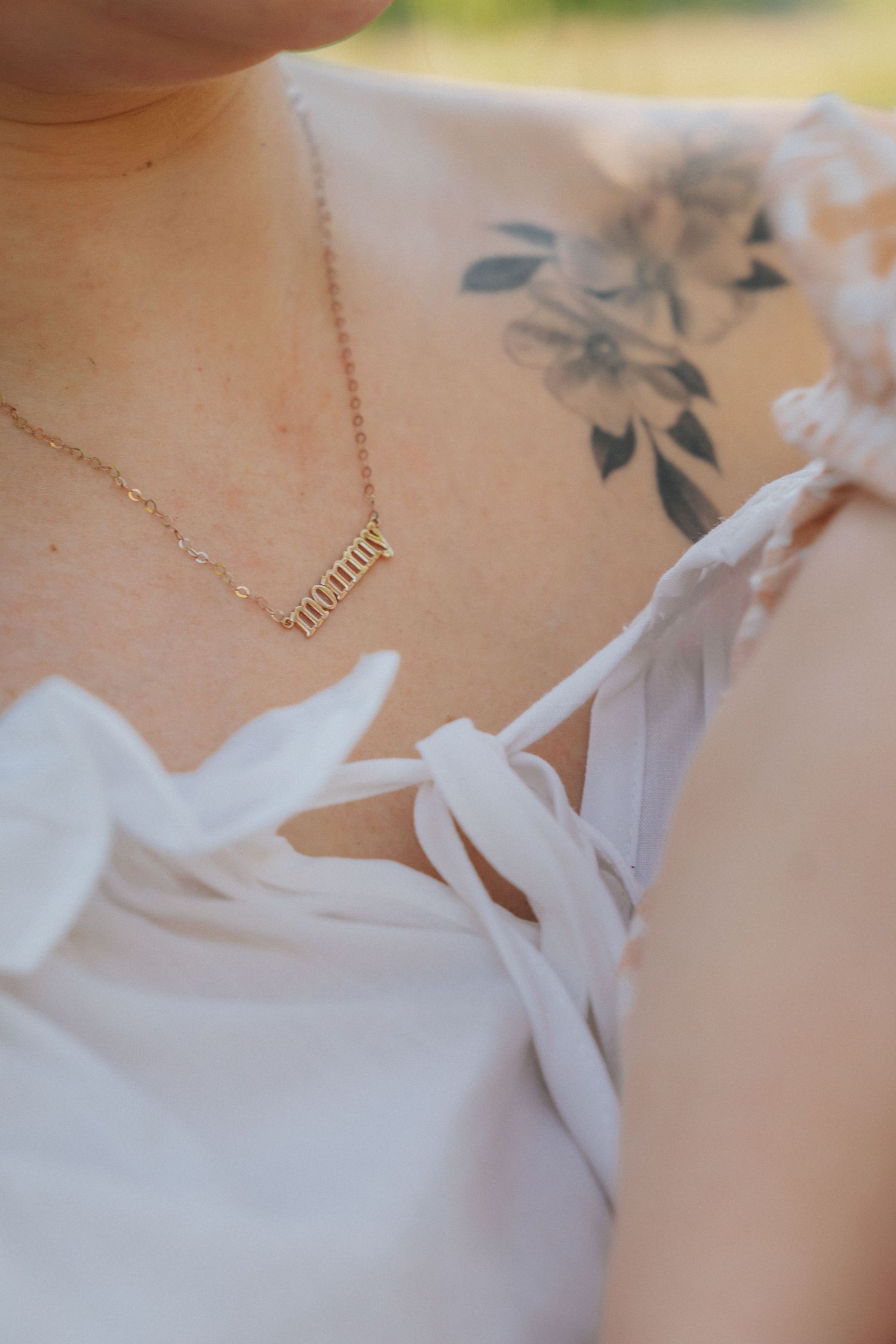 Gold mommy necklace on woman wearing white dress with floral shoulder tattoo.