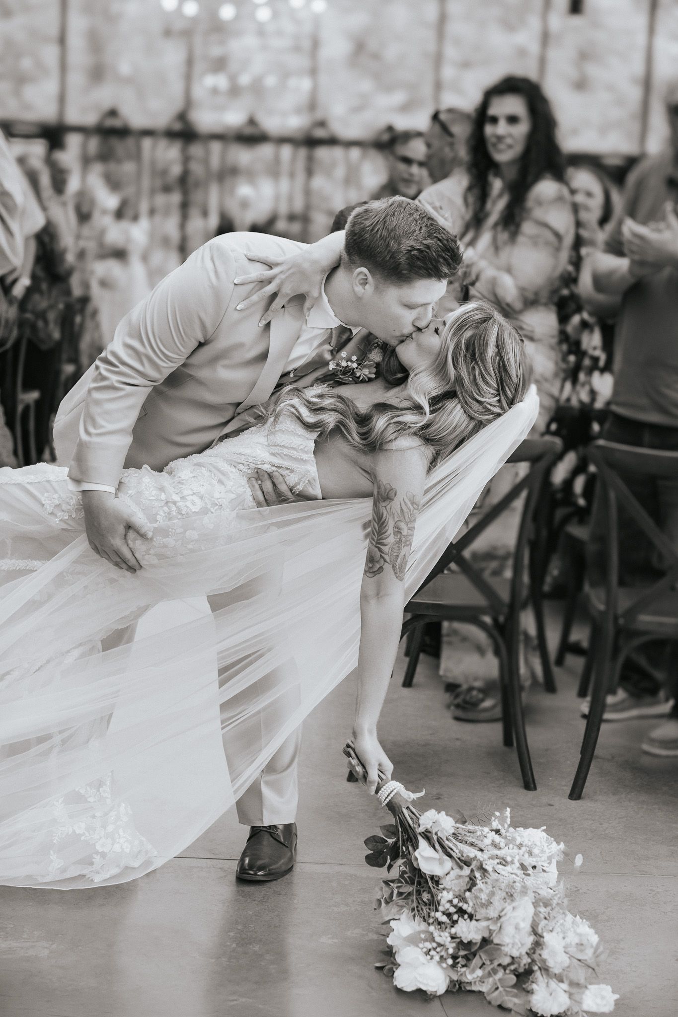 Black and white photo of groom dipping bride for a kiss at wedding ceremony while guests cheer in background.