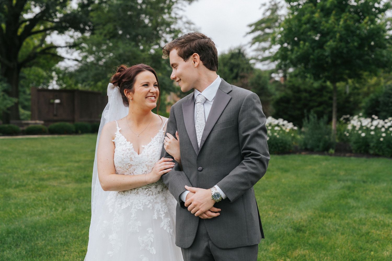 Bride in lace gown and groom in gray suit smile at each other in a lush garden setting.