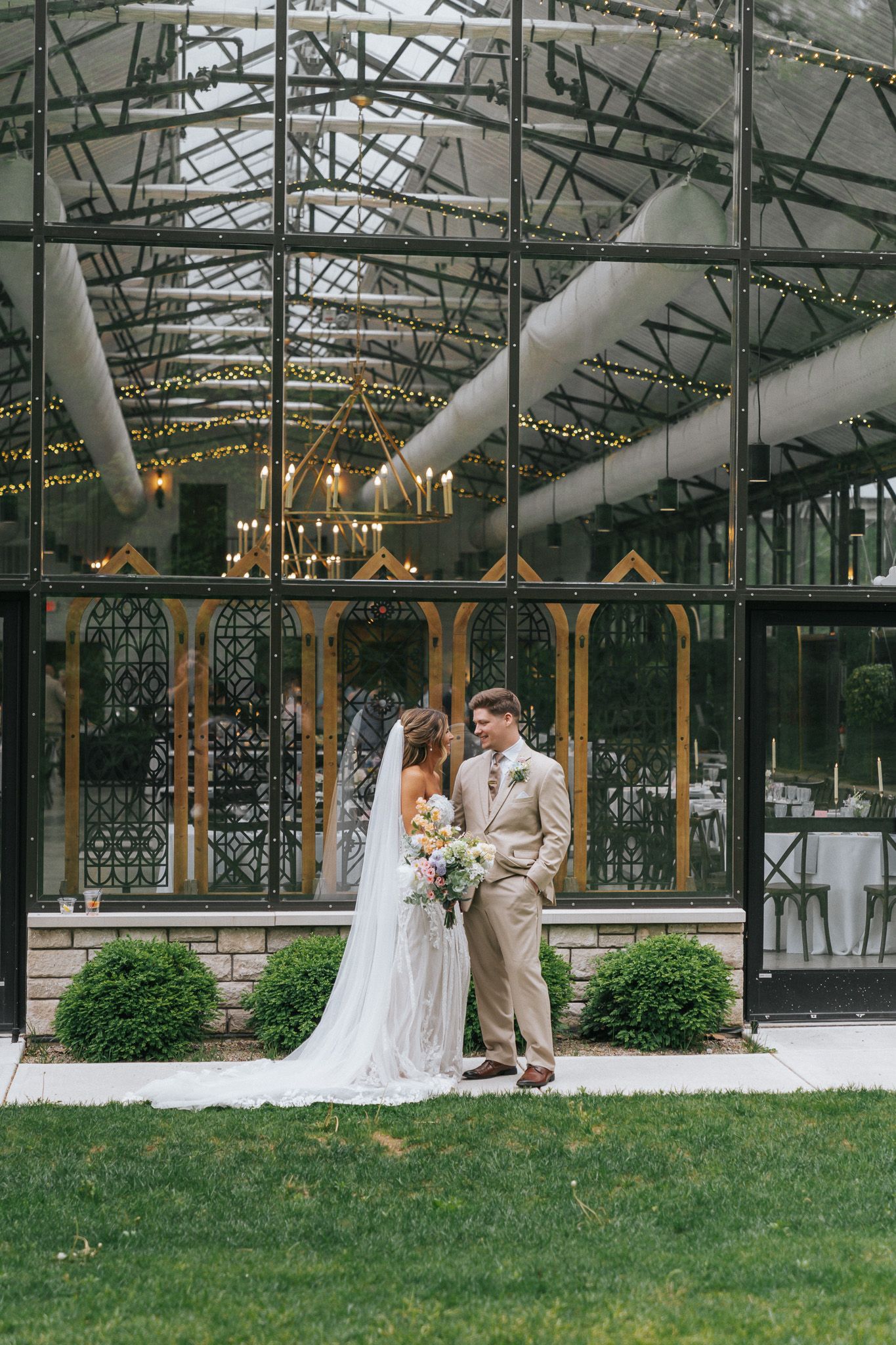 Bride and groom pose together outside an elegant glass greenhouse wedding venue with string lights and chandeliers.
