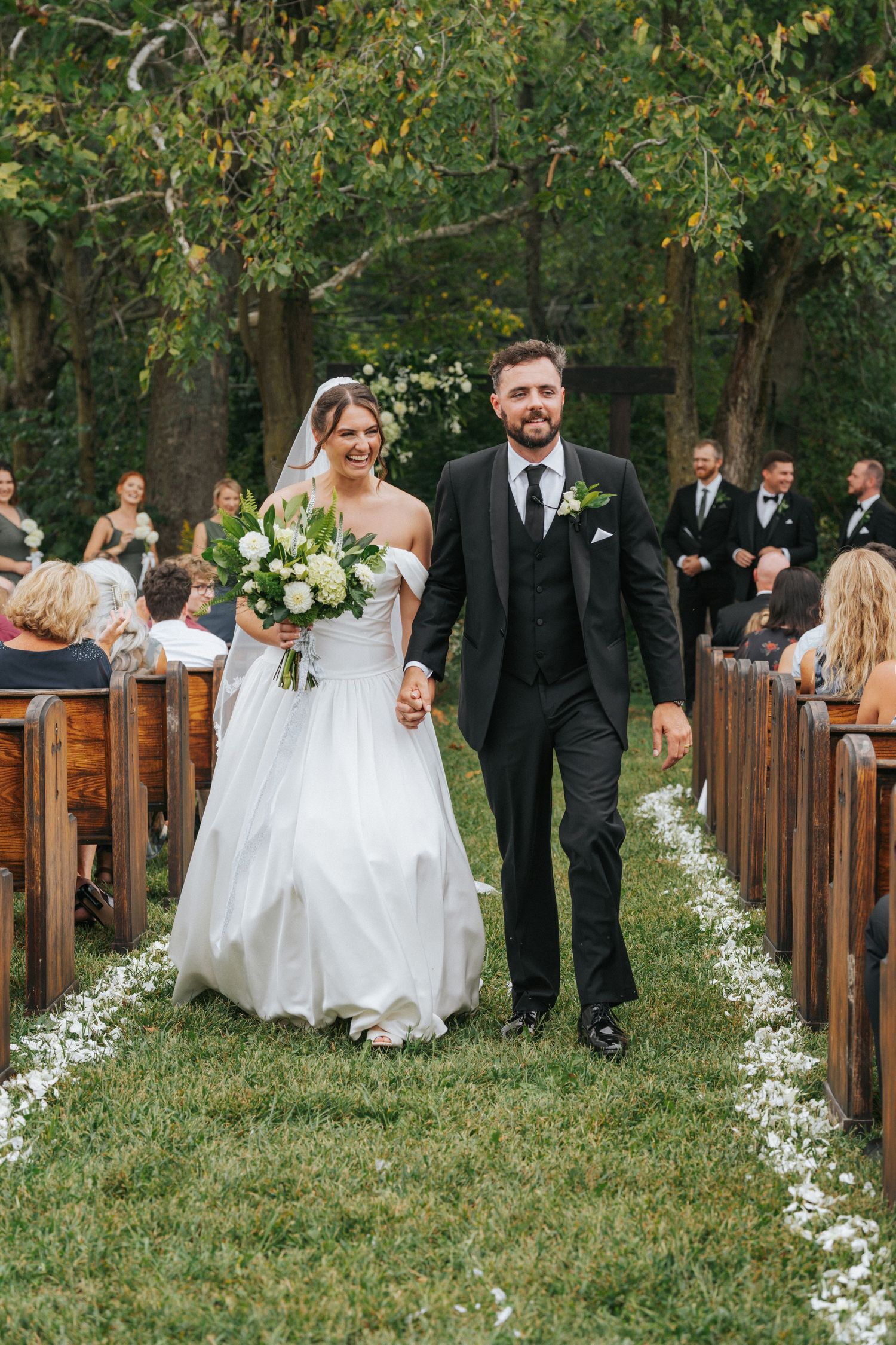 Bride and groom walk down outdoor aisle after wedding ceremony, surrounded by guests and lush greenery.
