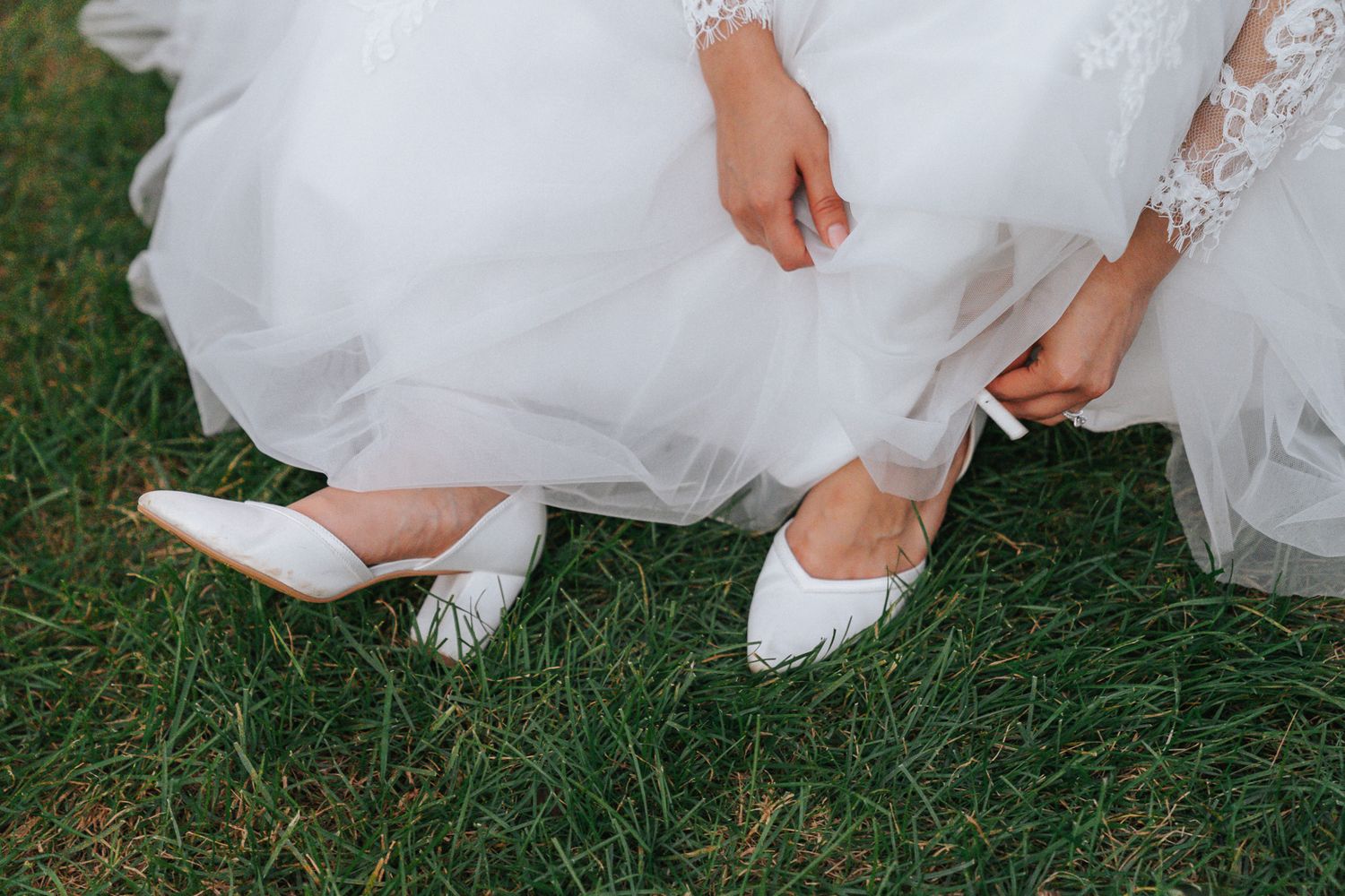 Bride in white lace wedding dress adjusting white pointed-toe bridal shoes while sitting on green grass.