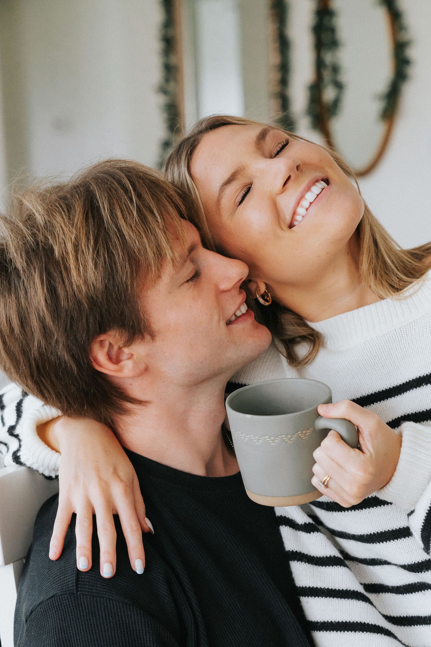 Happy woman in striped sweater holding a mug laughs as her partner kisses her cheek indoors.