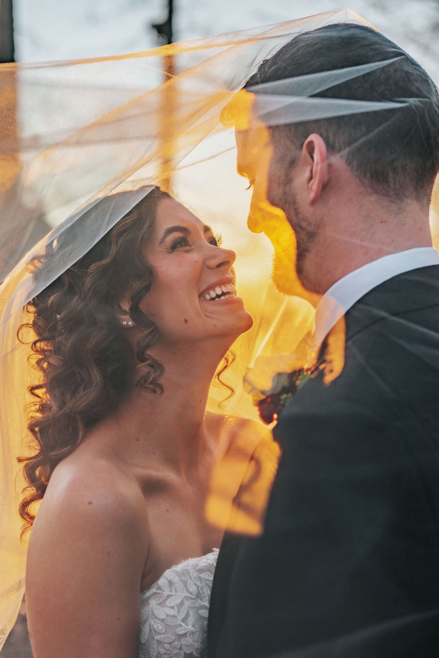 Bride laughs joyfully as groom holds her close under a veil glowing with warm golden sunset light.