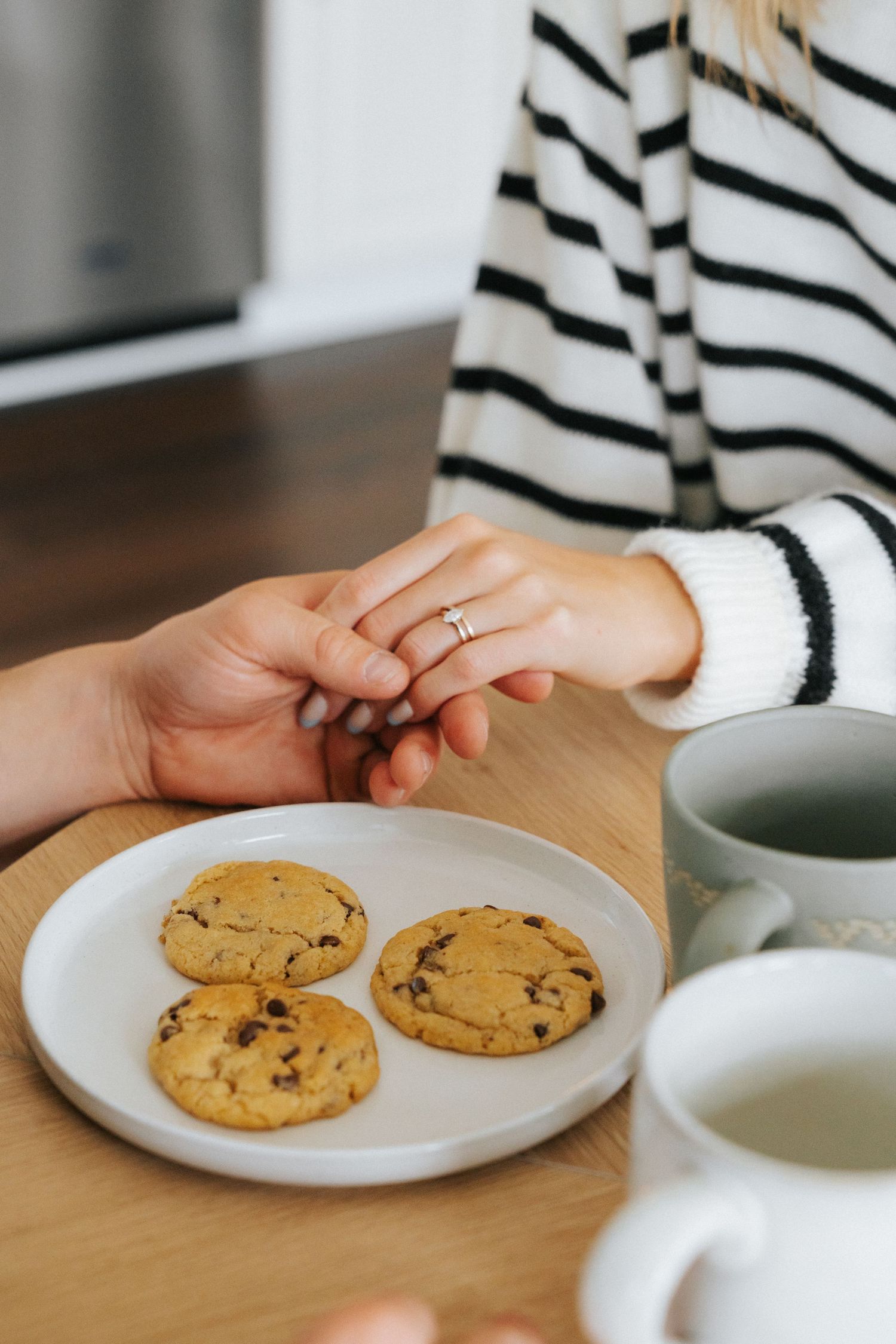 Couple holding hands over wooden table with chocolate chip cookies and mugs of tea or coffee.