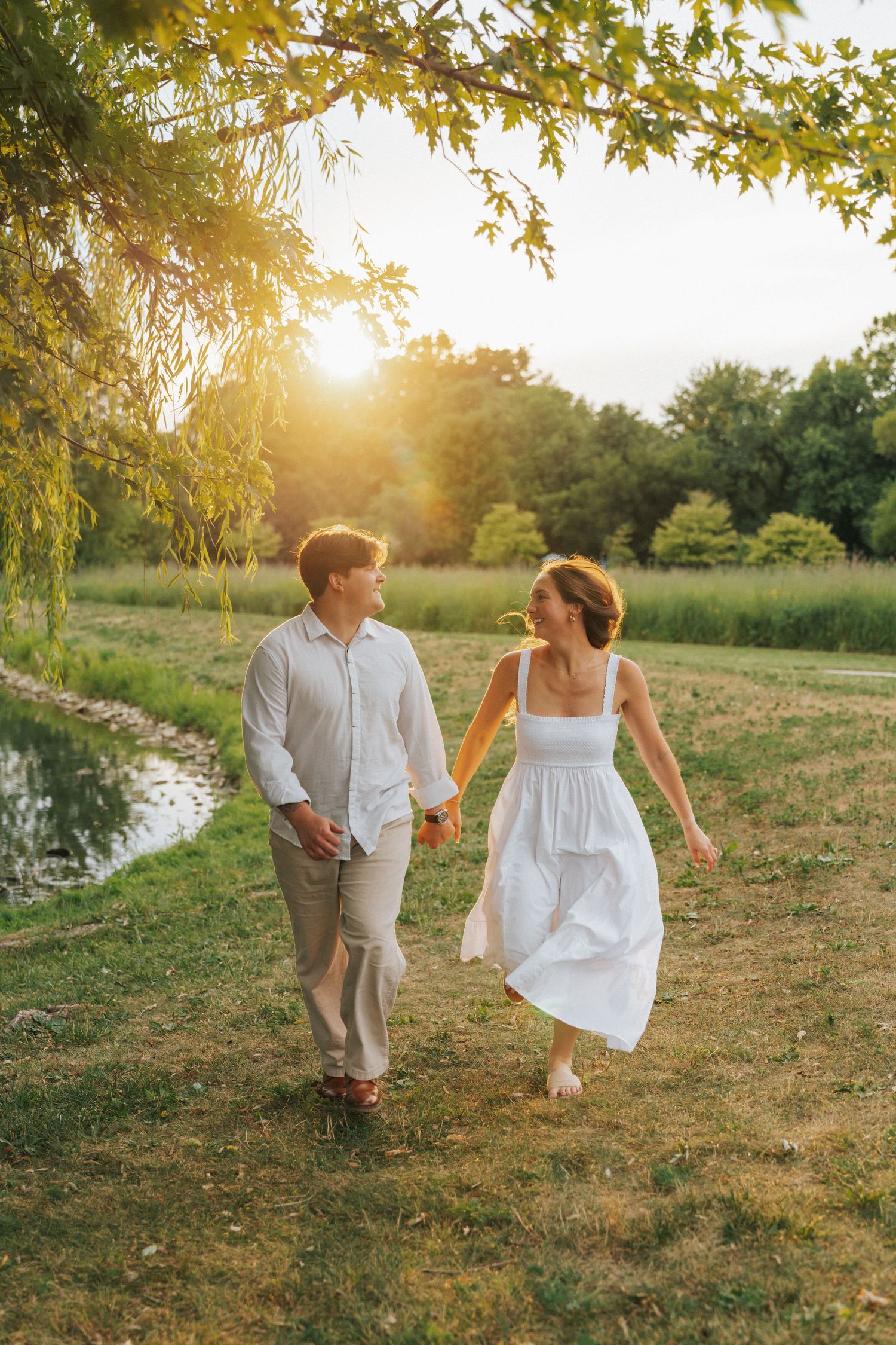 Couple holding hands walking in a sunny park near a pond, woman in white dress, man in white shirt, golden sunset light.
